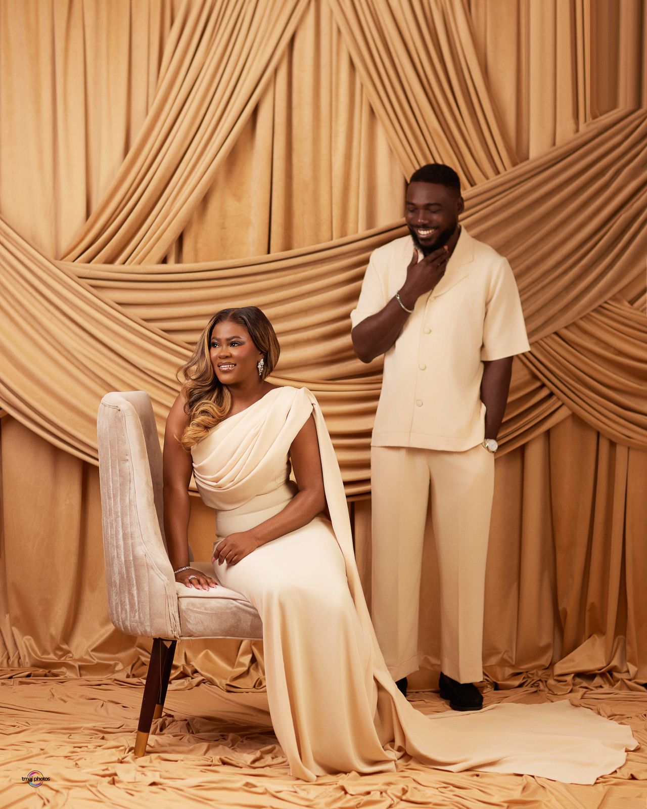 Elegant couple in matching cream outfits posing against draped gold fabric backdrop for a formal portrait shoot.