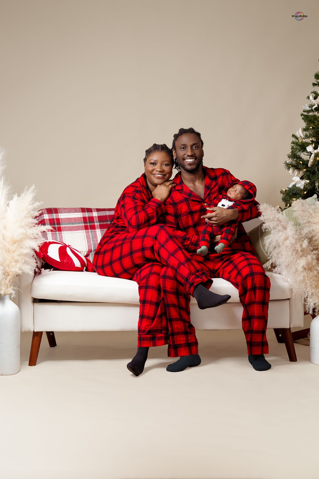 Family in matching red plaid pajamas on white sofa, father holding newborn baby near Christmas tree.