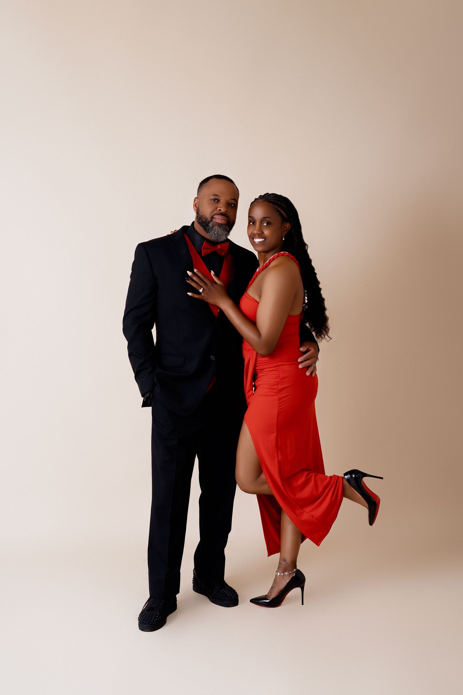Elegant couple posing in studio, man in black tuxedo with red bow tie, woman in red slit dress and heels.