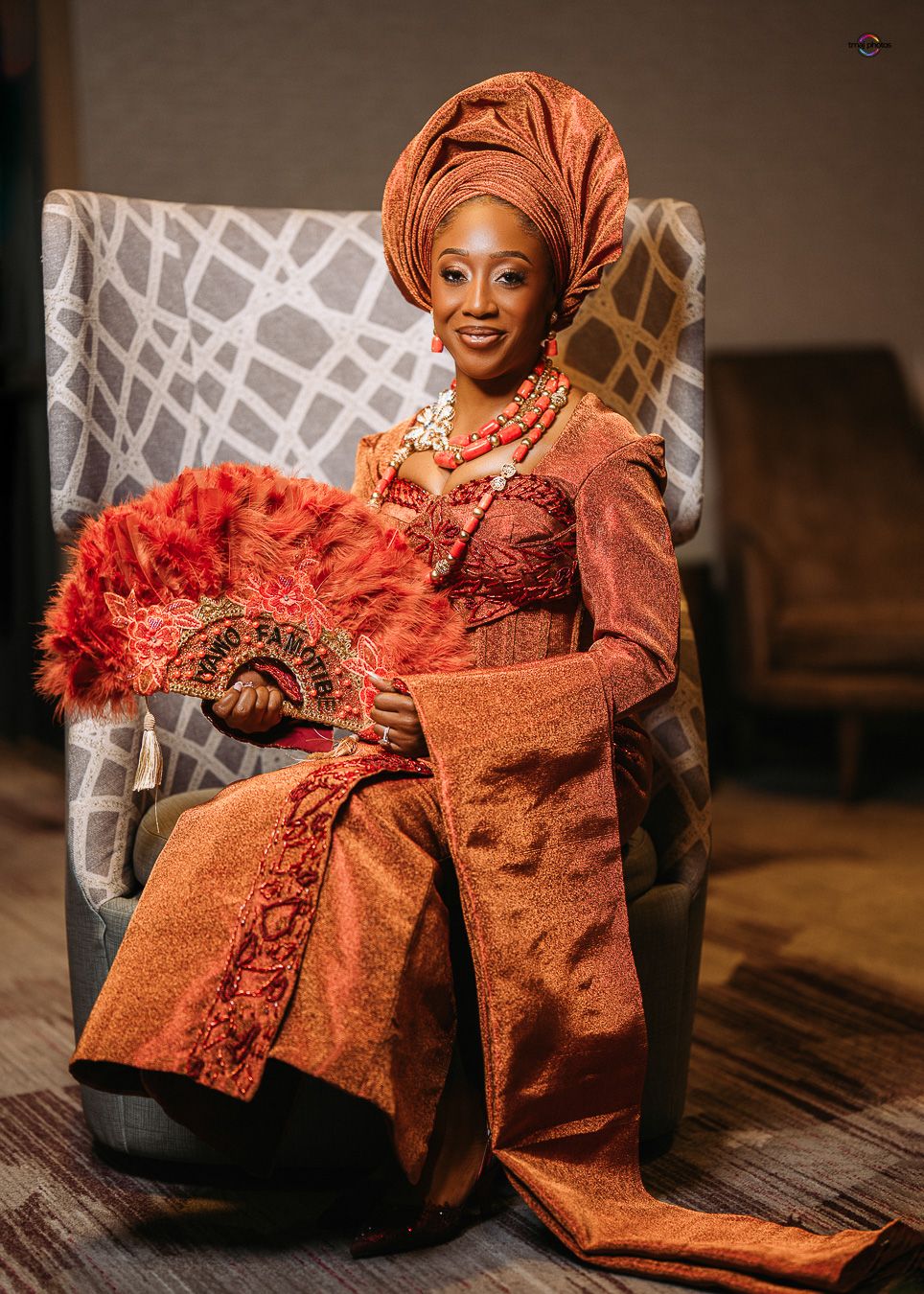 Woman in traditional Nigerian attire with orange gele headwrap, aso-oke outfit, coral beads, and feather fan seated in chair.