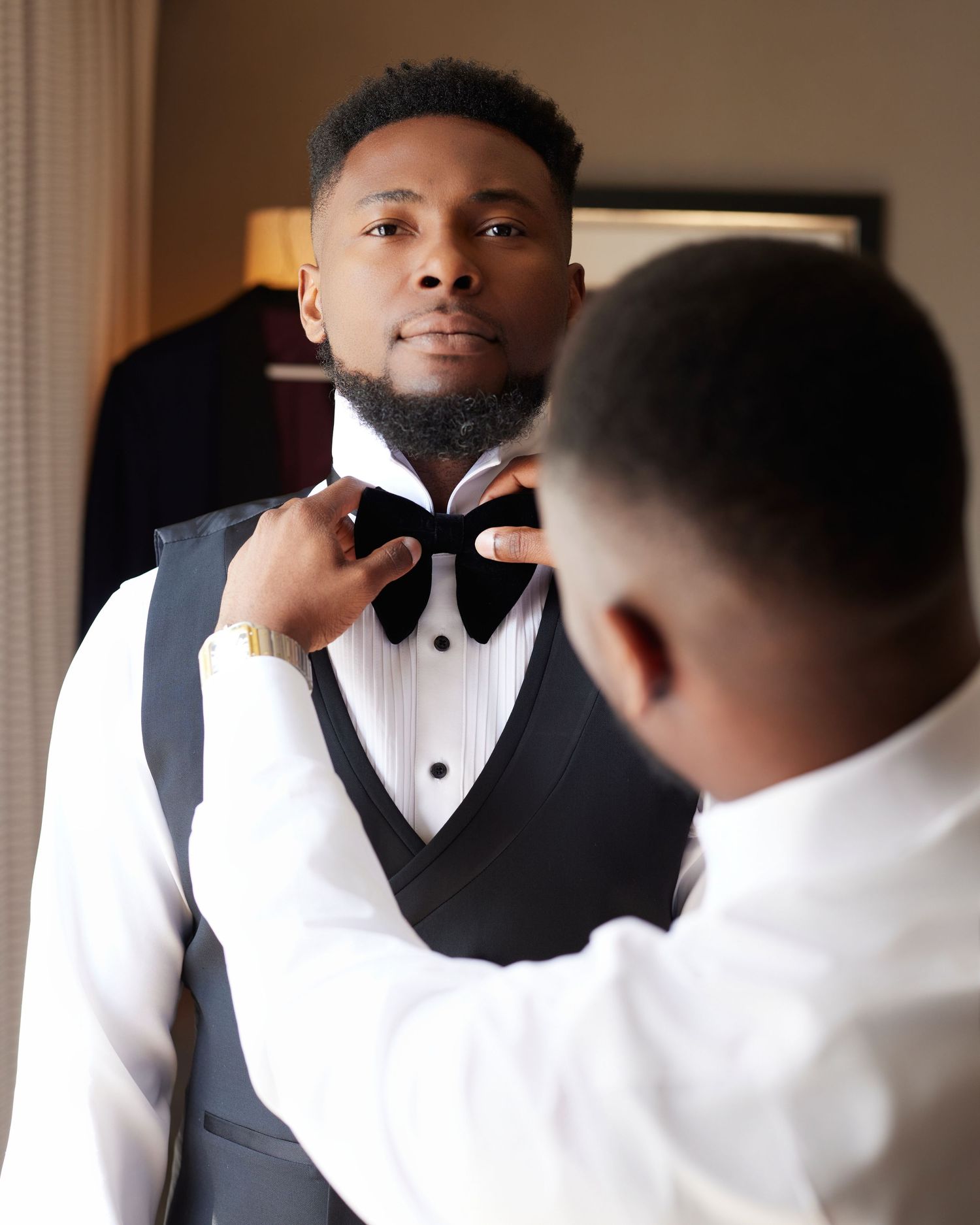 A man in formal attire gets his black bow tie adjusted by another person while preparing for a special event.
