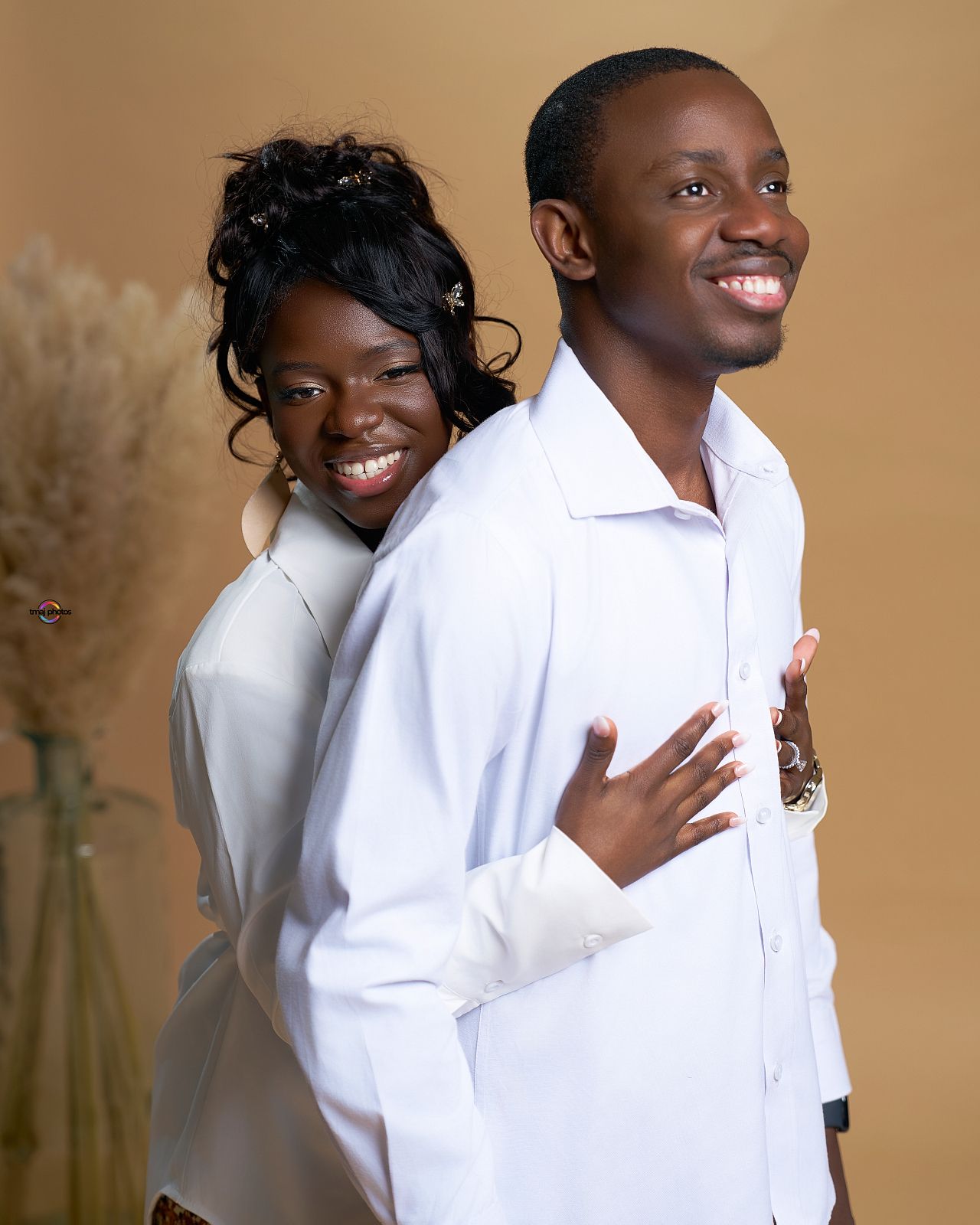 A smiling couple in matching white shirts pose back-to-back in a studio portrait with a warm beige background.
