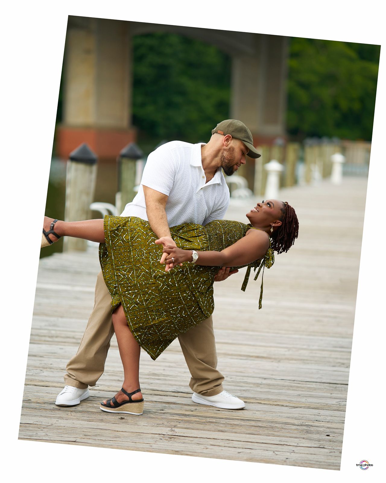 Couple dancing on a wooden dock, man dipping woman in a green patterned skirt, with a bridge in the background.