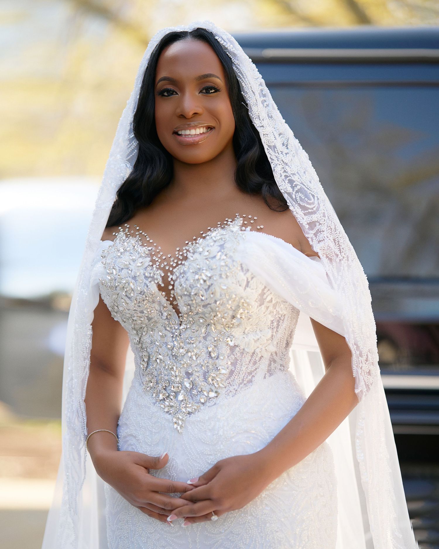 Bride in a beaded off-shoulder wedding gown with a lace cathedral veil, smiling outdoors.