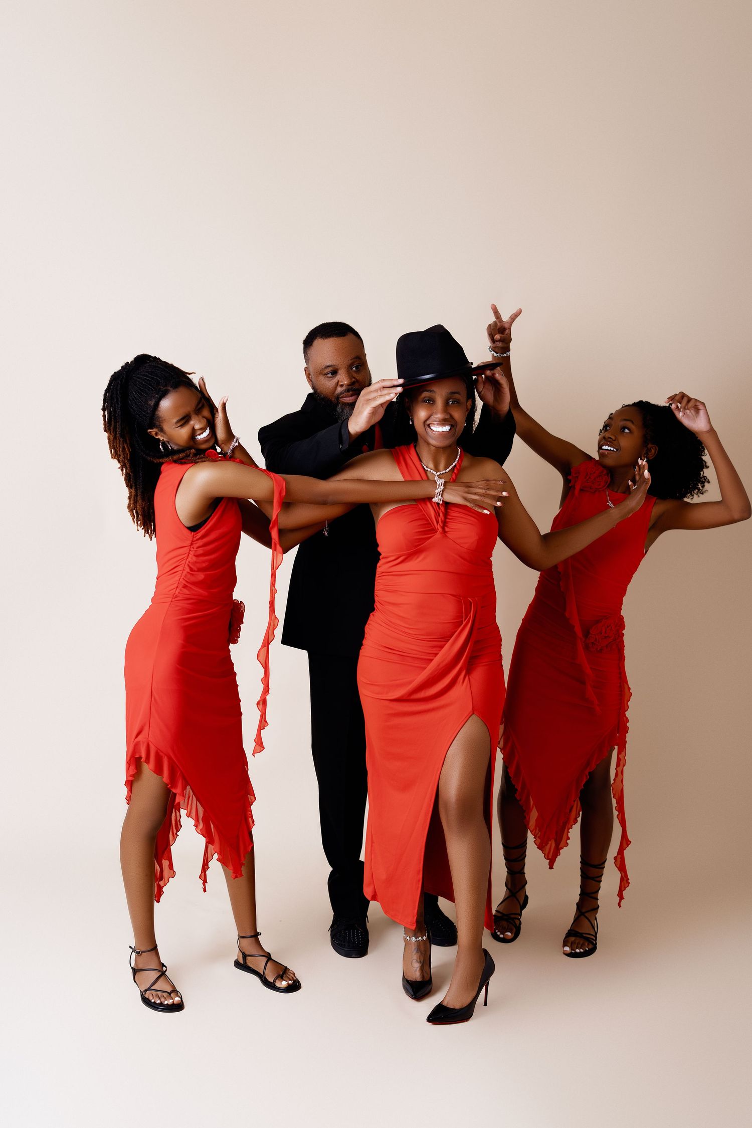 Four people posing in studio, man in black suit with three women in matching red dresses and a fedora hat.