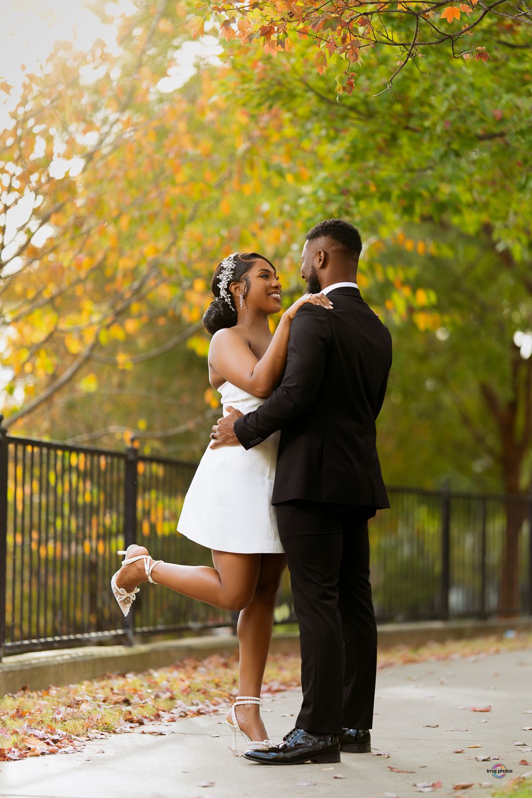 Bride and groom embrace joyfully outdoors among colorful autumn foliage, bride lifting one leg in white dress.