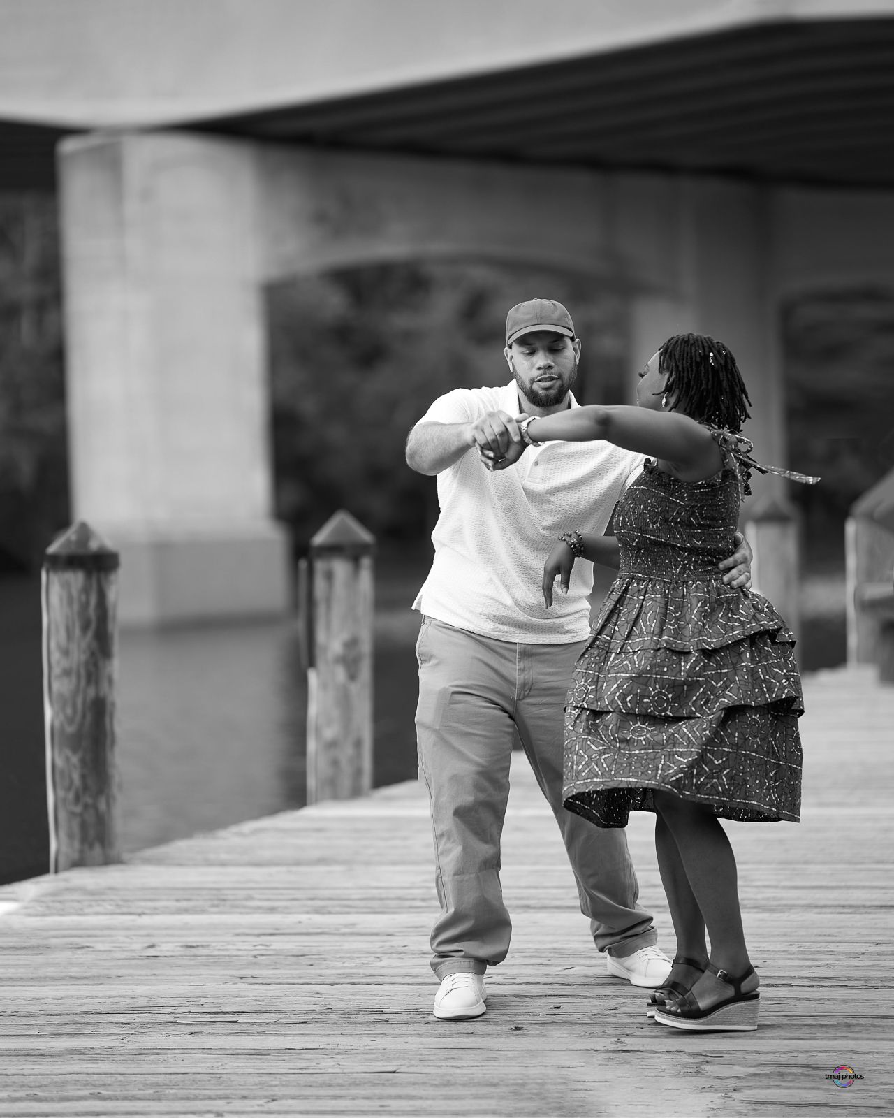 Black and white photo of a couple dancing on a wooden dock near a waterfront.