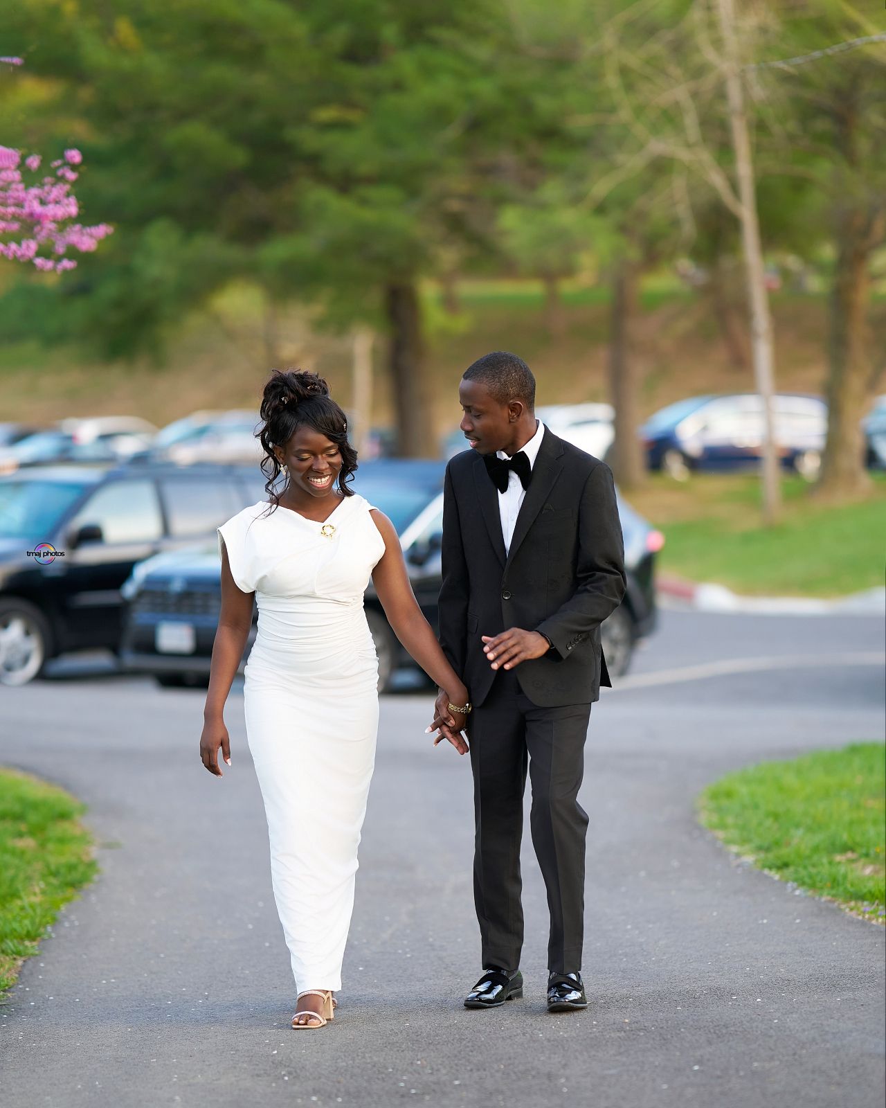 A couple in formal attire walks hand-in-hand on a path, woman in white dress, man in black tuxedo, trees in background.