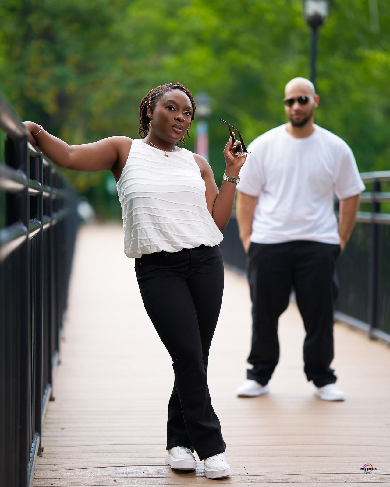 A woman in white top and black flare pants poses on a wooden bridge walkway, with a man in white standing behind her.