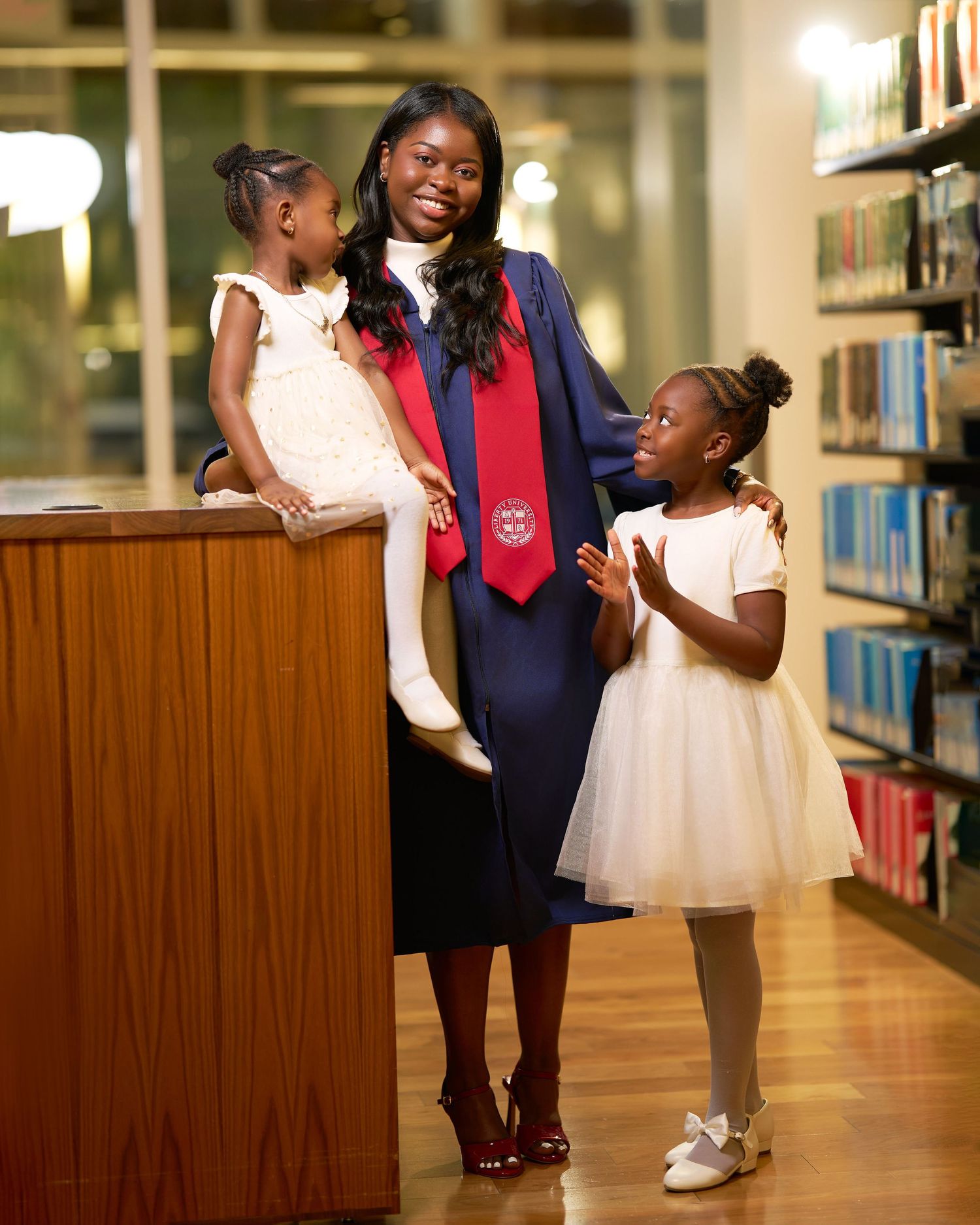 Graduate in cap and gown smiles with two young girls in white dresses inside a library.