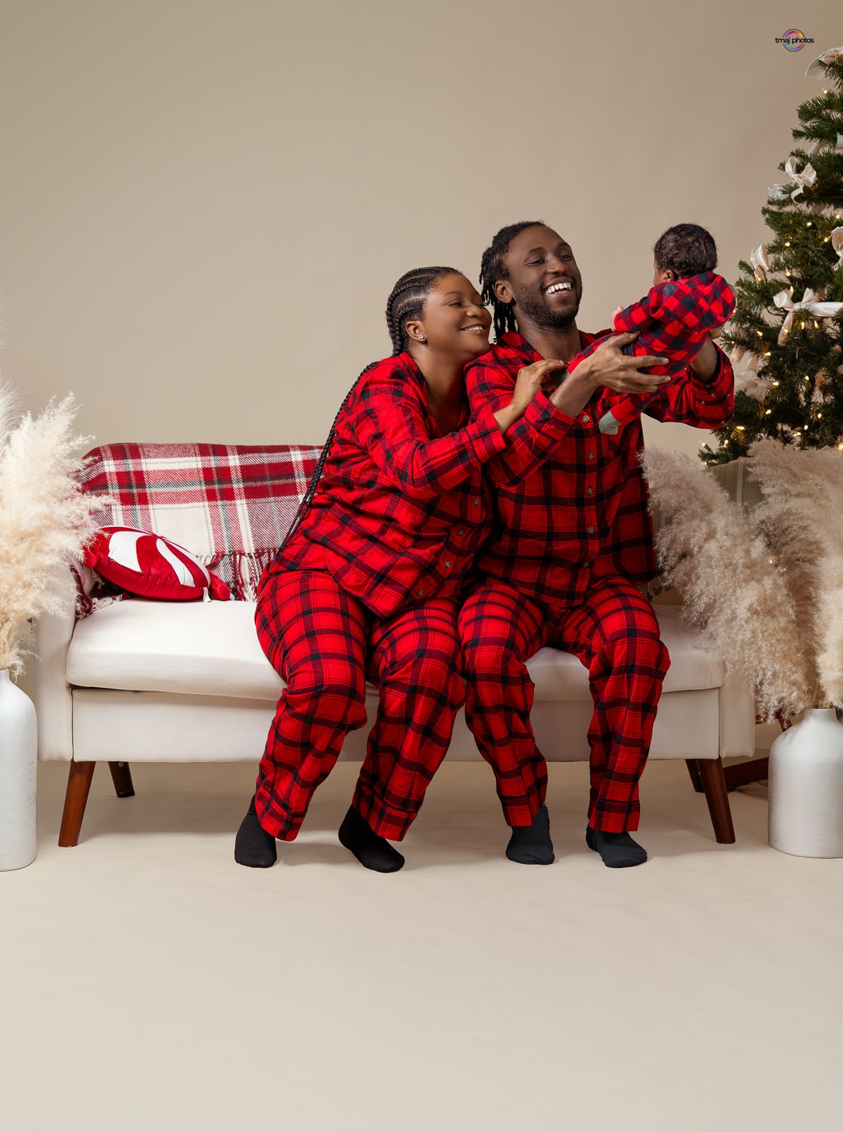 Happy family in matching red plaid pajamas on sofa, parents lifting smiling baby near Christmas tree.