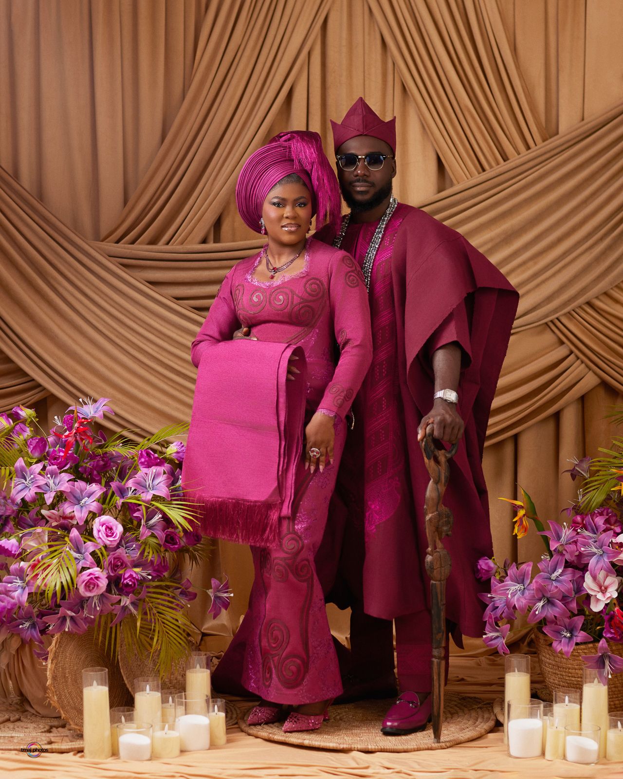 Couple in matching magenta Nigerian traditional attire with gele headwrap, posing among purple flowers and candles.