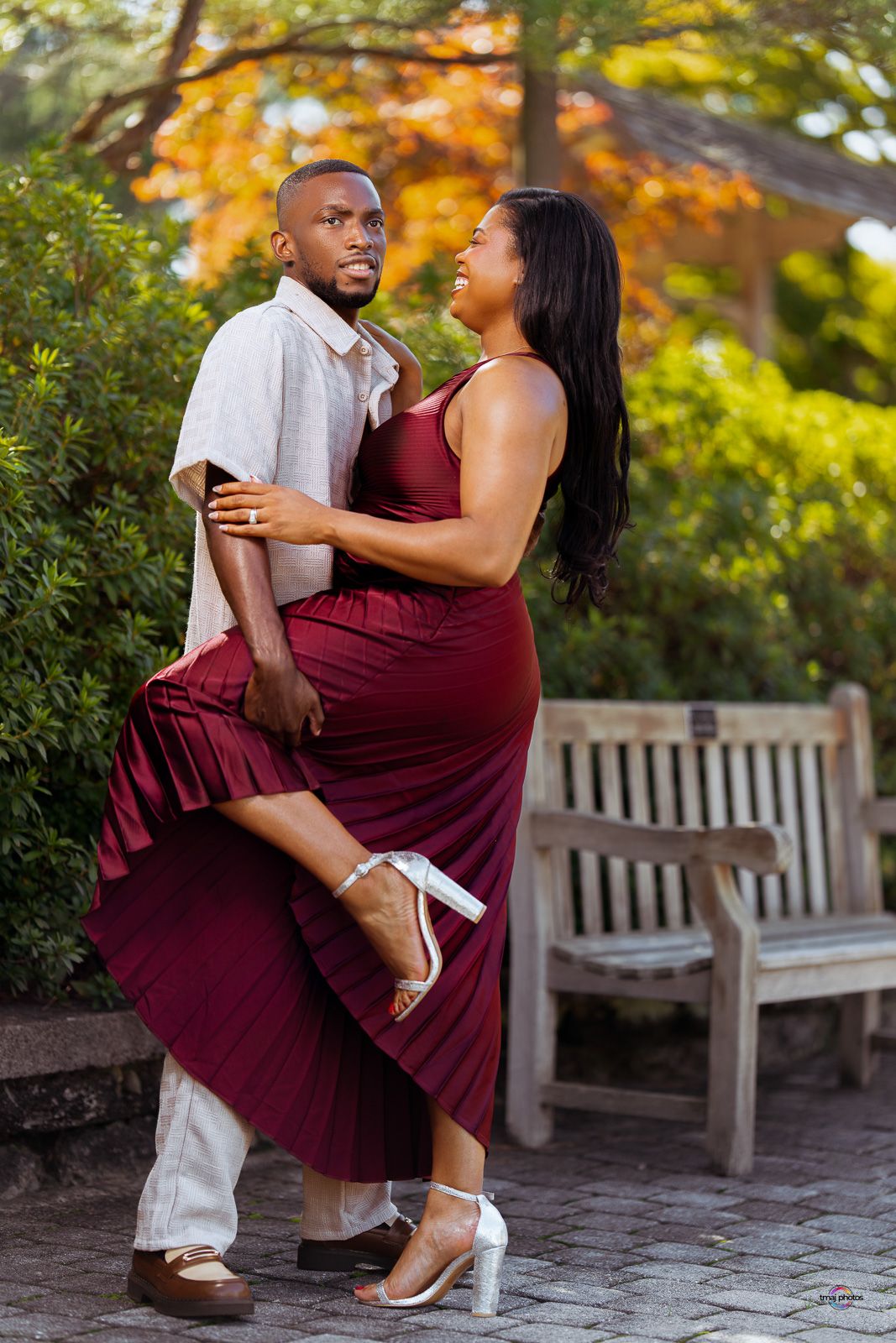 Couple embraces romantically in garden setting, woman in burgundy dress lifts leg while laughing with partner.