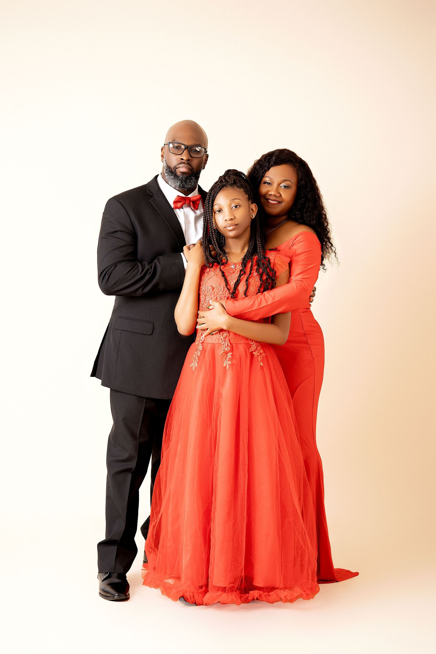 A family of three poses in coordinating red outfits, with a man in a tuxedo and two women in elegant red gowns.