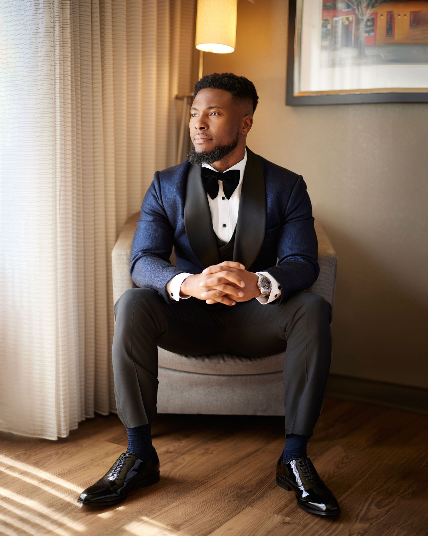 Elegant man in navy tuxedo with bow tie seated in armchair, exuding sophistication in formal attire.