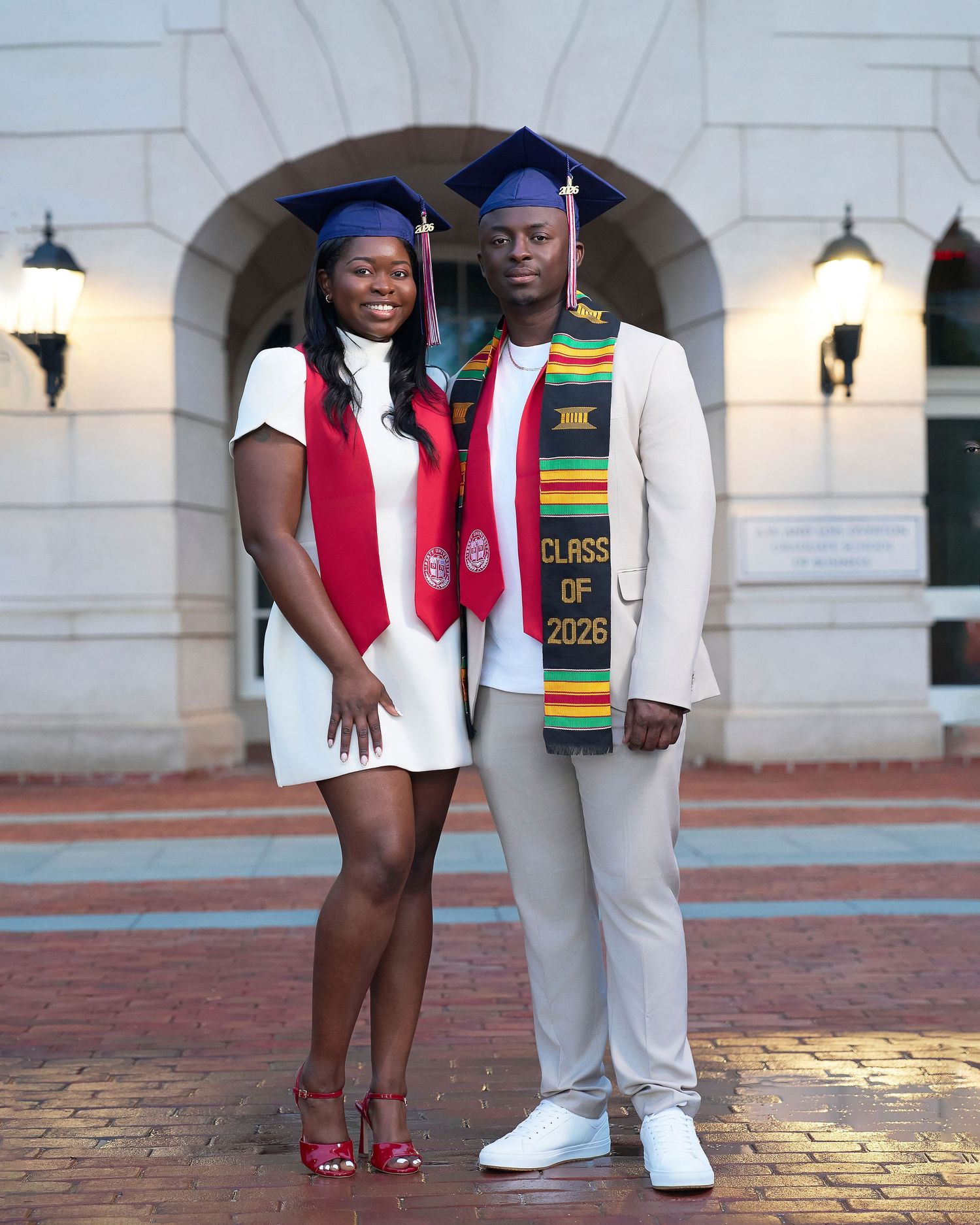 Two graduates in blue caps and white outfits with colorful stoles pose proudly for graduation photos.