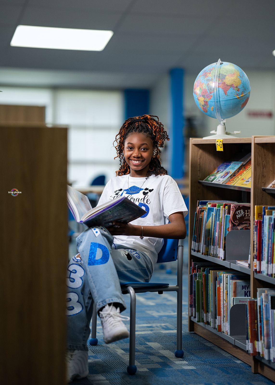 Smiling student reading a book in a school library with a globe and colorful bookshelves in the background.