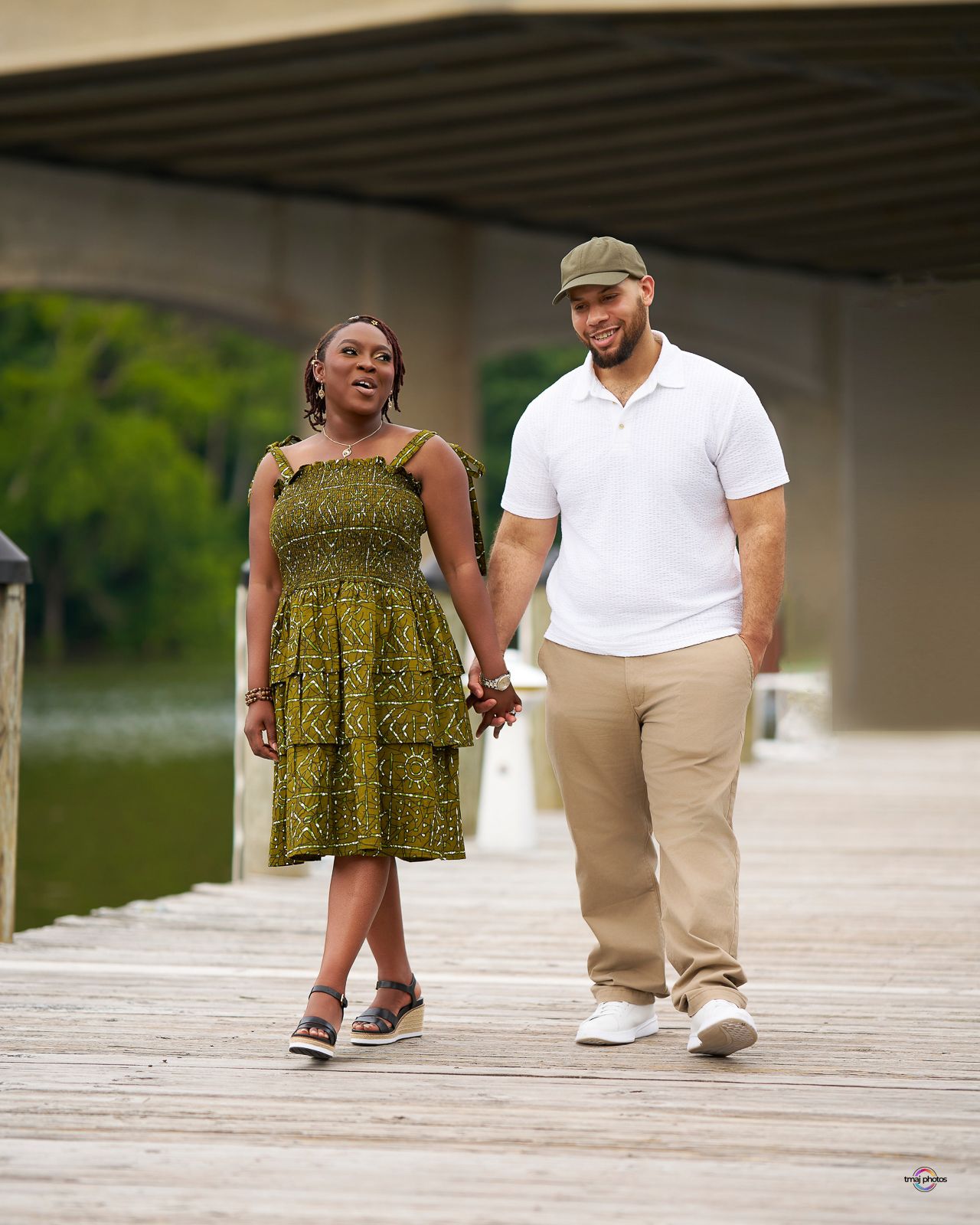 Couple holding hands walking on a wooden dock near water, woman in green dress, man in white polo and khaki pants.