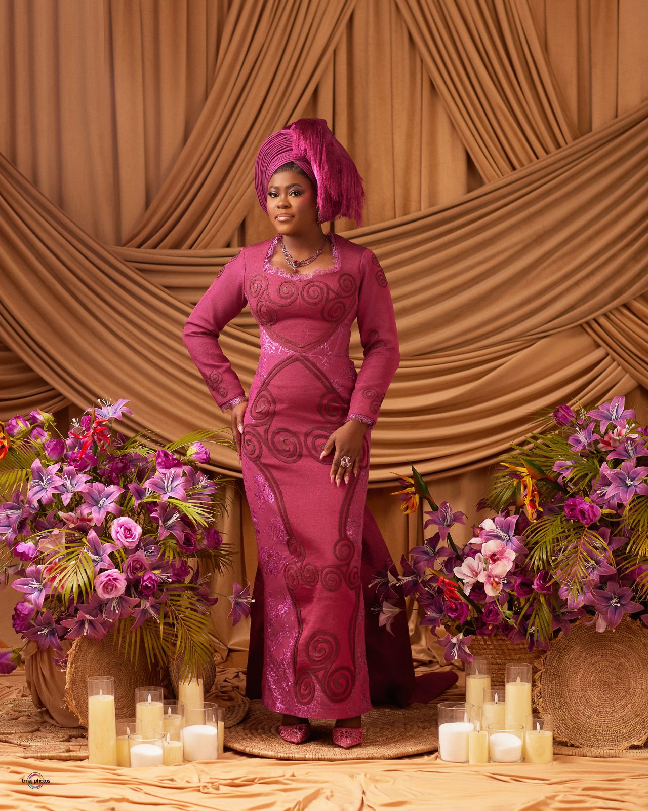 Woman in elegant magenta Nigerian traditional attire and gele headwrap poses before draped backdrop with floral arrangements.