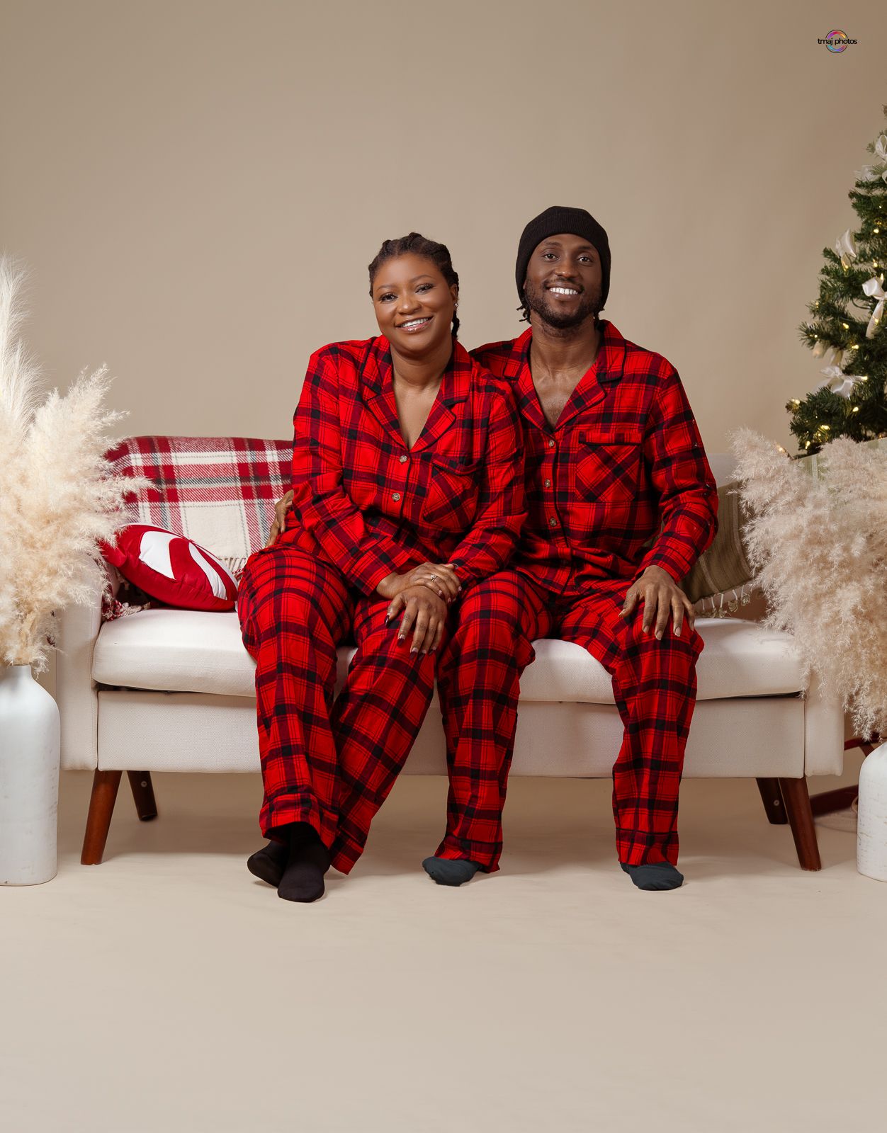 Couple wearing matching red plaid pajamas sitting on white sofa with Christmas tree in background.