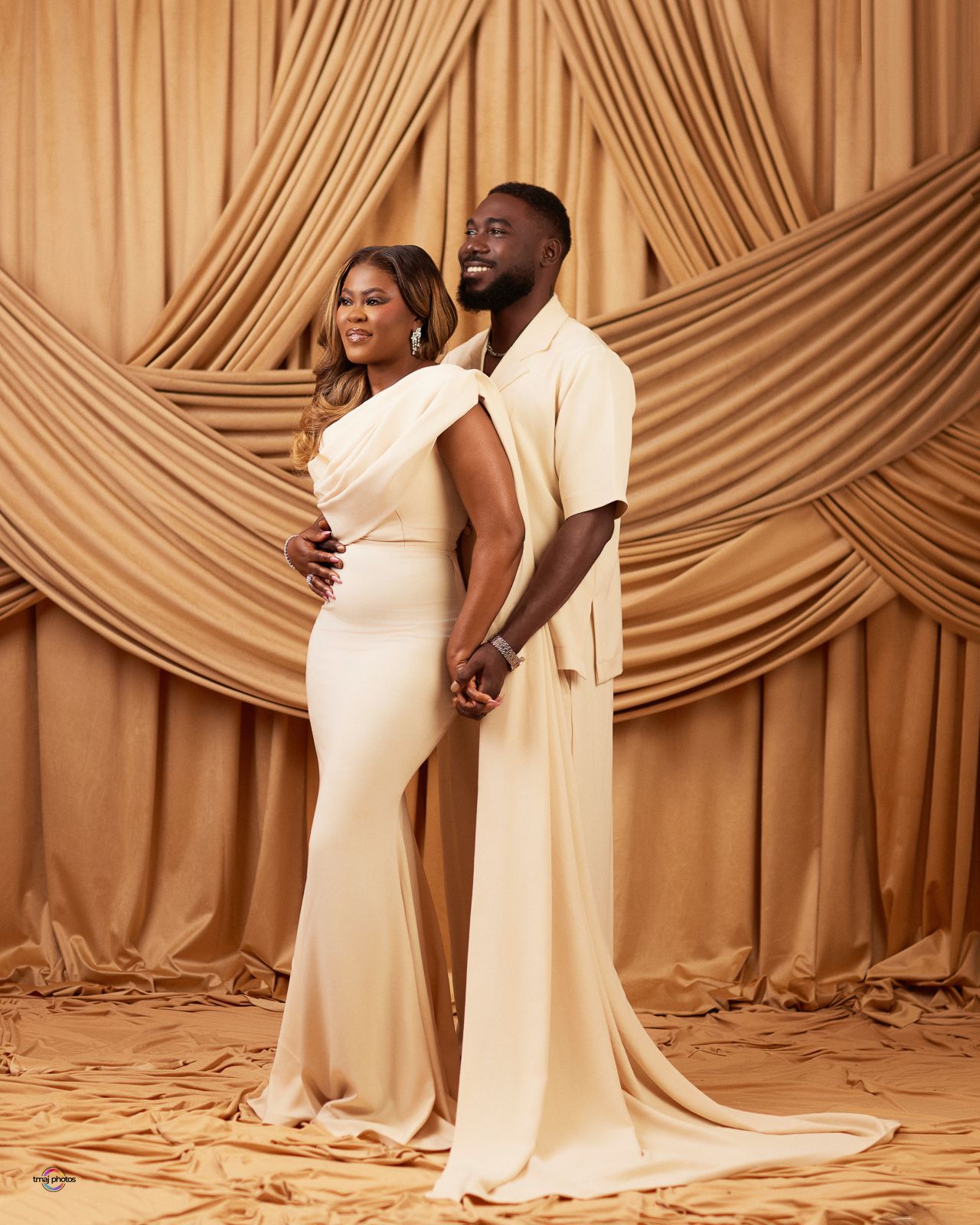 Elegant couple in matching cream outfits posing before draped golden fabric backdrop in a maternity photoshoot.
