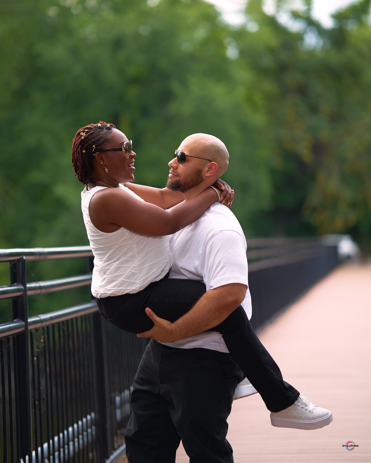 A man lifts a woman in his arms on a bridge, both smiling and wearing white tops with black pants.