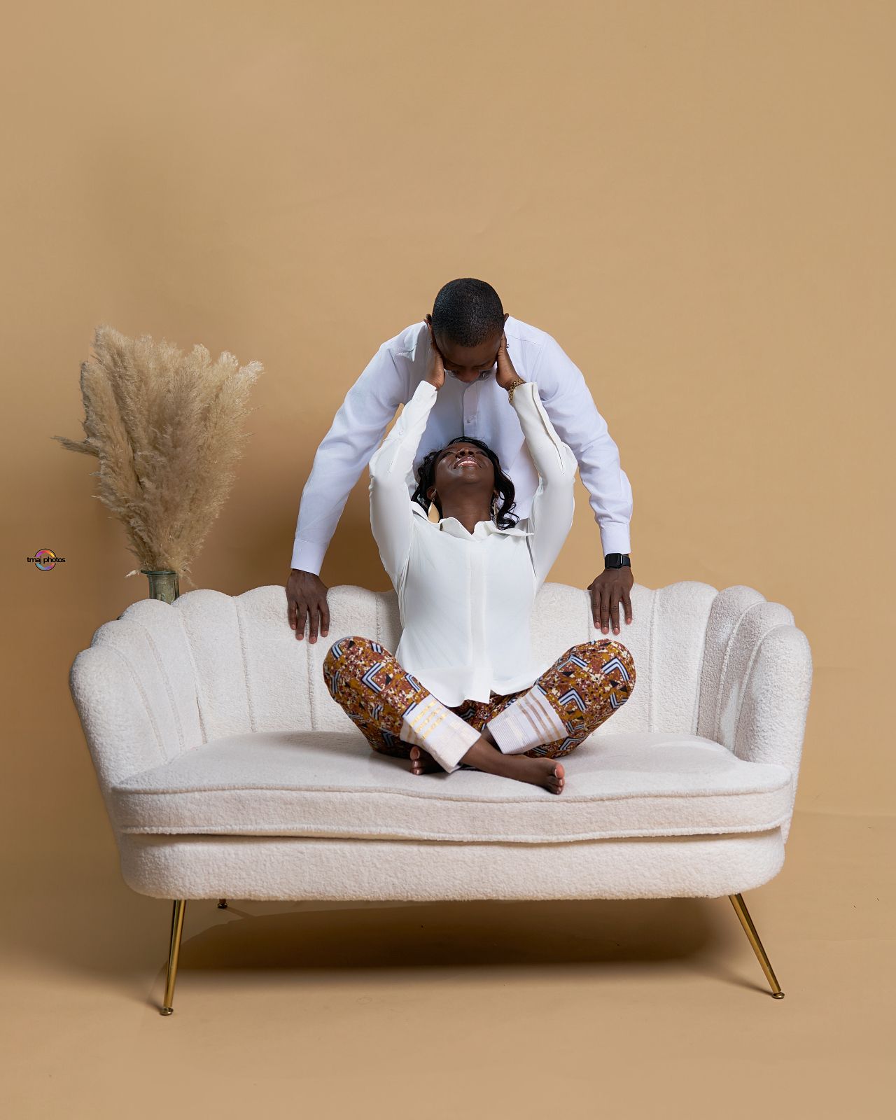A couple poses on a cream velvet sofa, woman sitting cross-legged in white outfit while man stands behind her on beige backdrop.