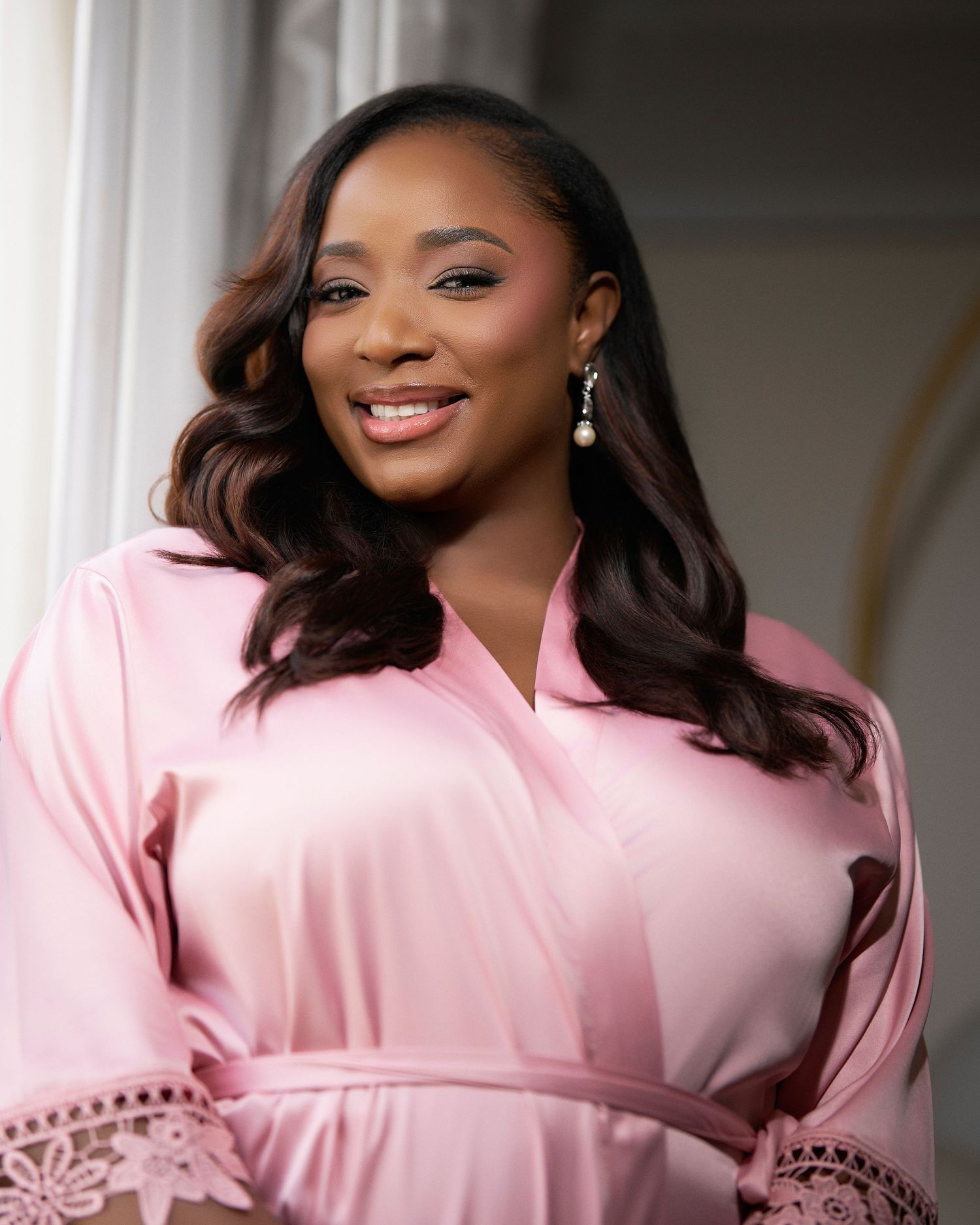 Smiling woman in pink satin robe with pearl earrings and wavy brown hair in bridal getting ready photo.