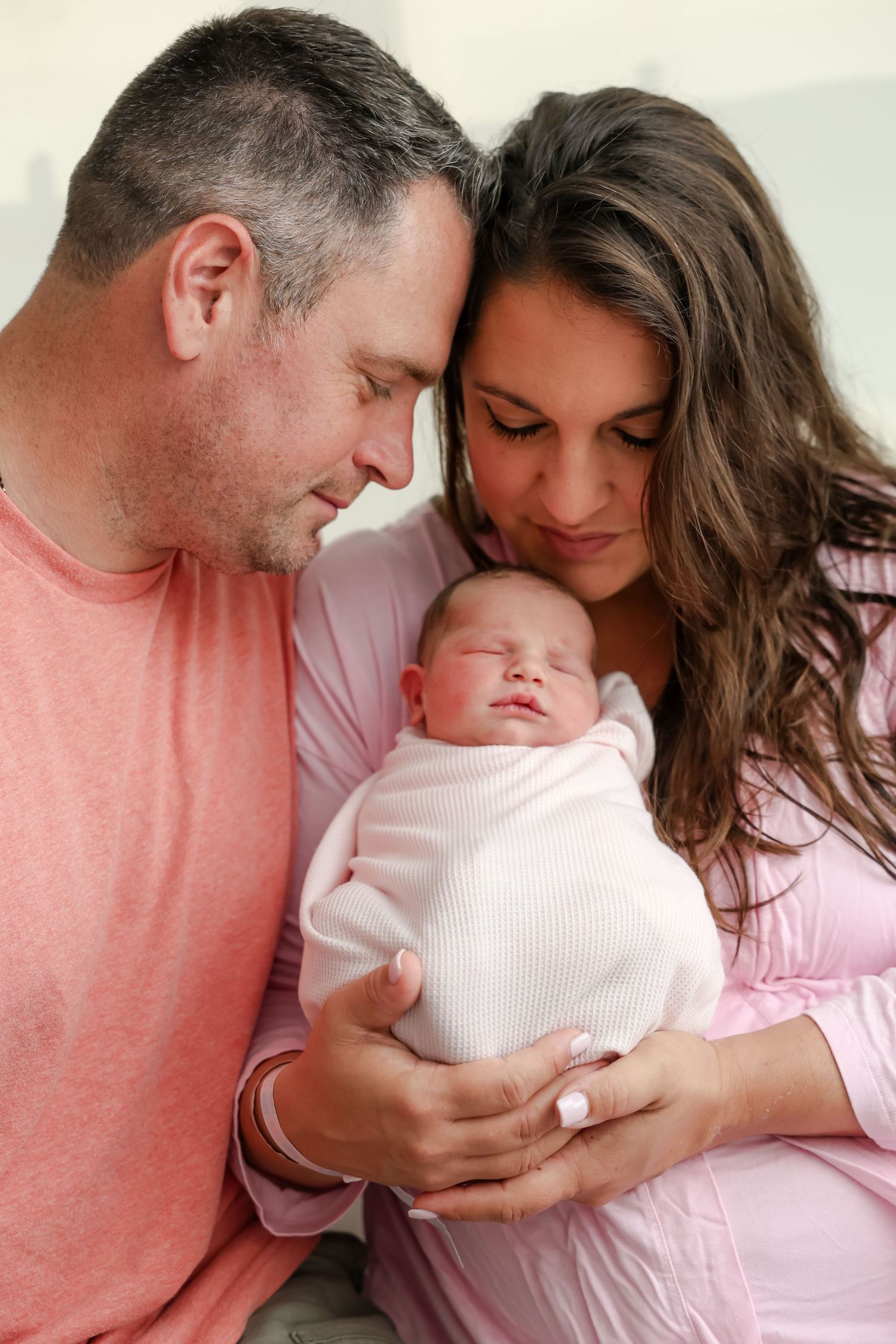 New parents tenderly hold their sleeping newborn baby wrapped in white, sharing an intimate family moment.