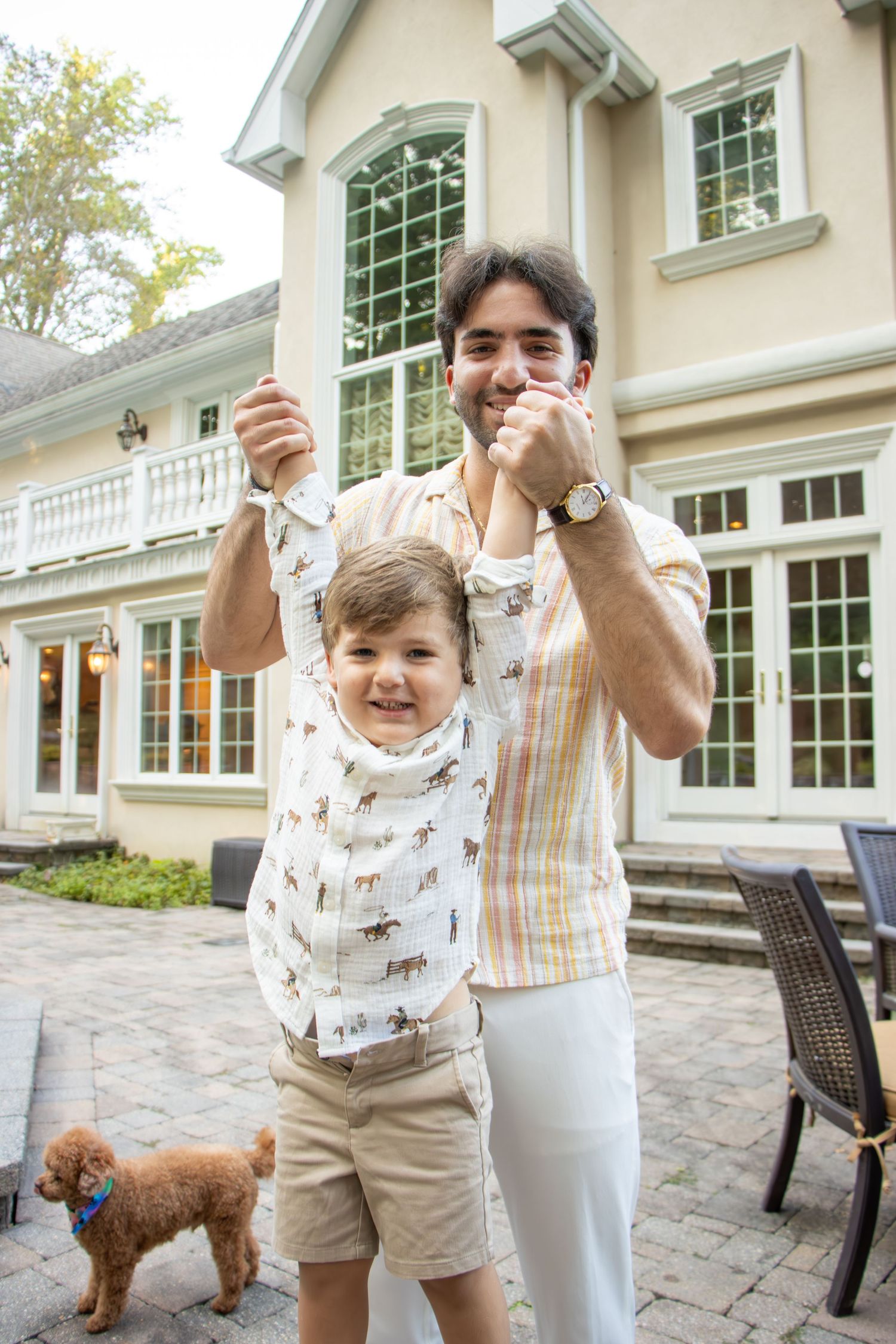 Man lifting smiling toddler boy by hands in backyard patio of large home with small brown dog nearby.