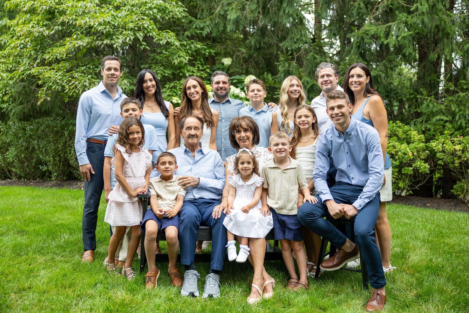 Large multigenerational family posing together outdoors on green lawn with lush trees in background.