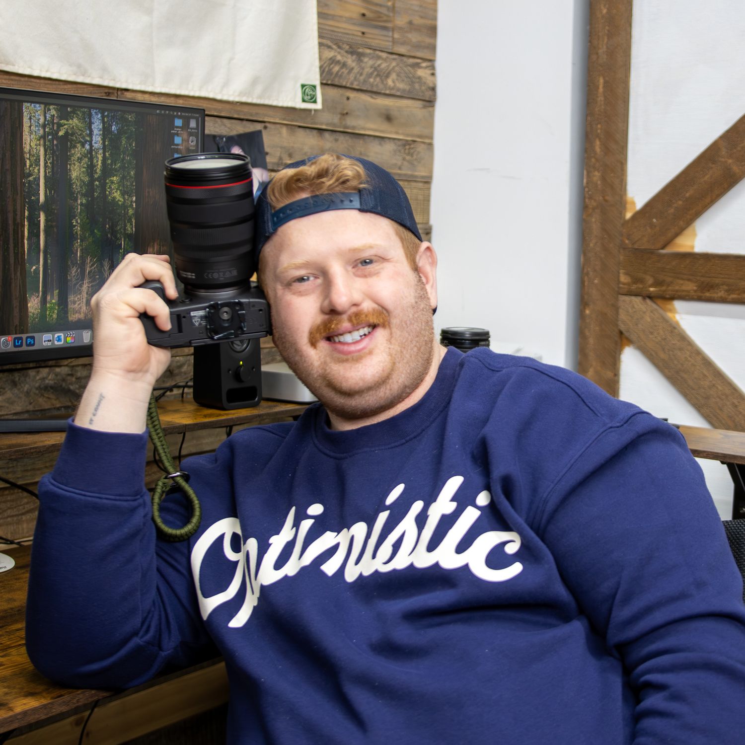 Smiling photographer holds a Canon camera in a rustic studio wearing an Optimistic sweatshirt.
by Andrew Fein Photography - Family and Newbo