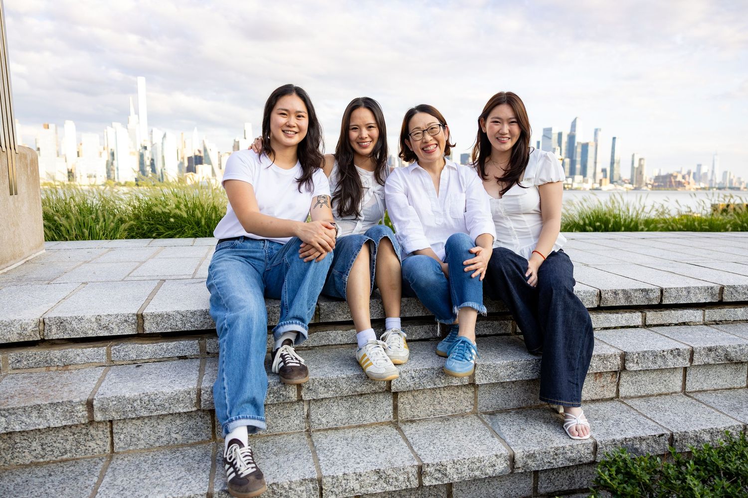 Four young women in white tops and jeans sitting on stone steps with a city skyline in the background.