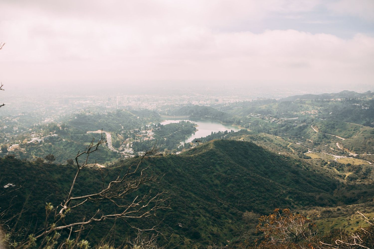 Aerial view of lush green hills overlooking a misty city valley with a reservoir on a cloudy day.
