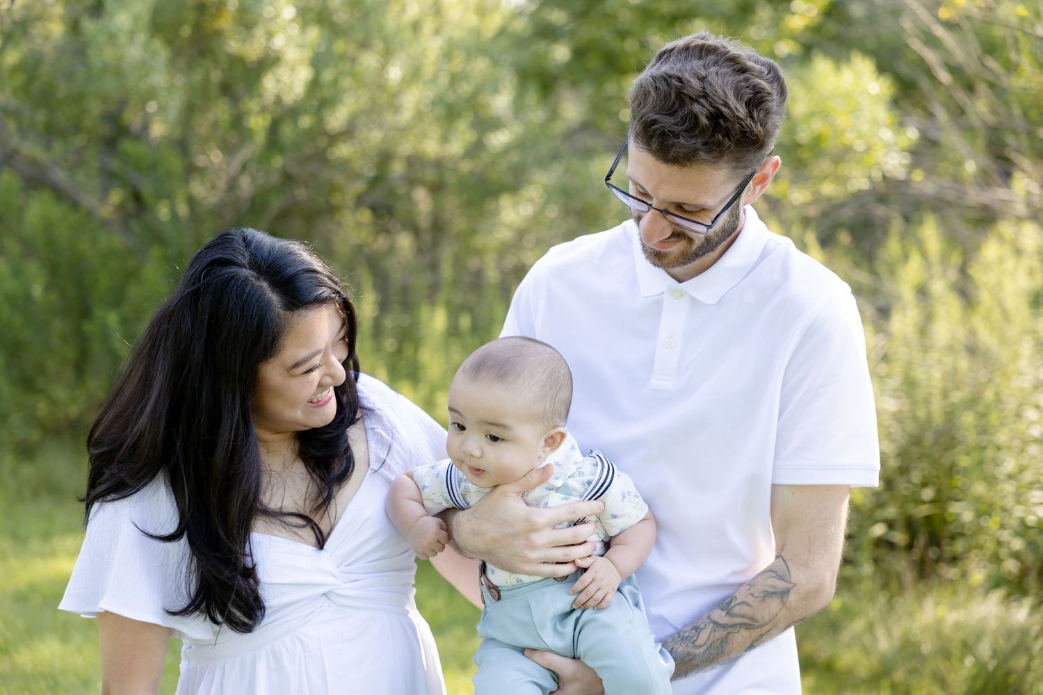 A young family with a baby poses outdoors in a lush green park, wearing white and light blue outfits.