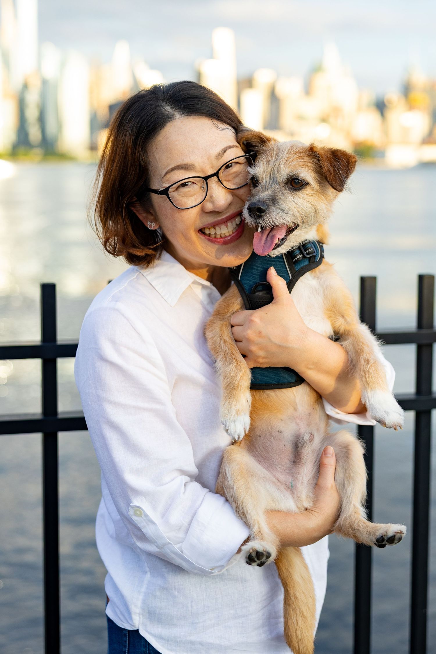 Smiling Asian woman holding a happy terrier mix dog with NYC skyline in background.