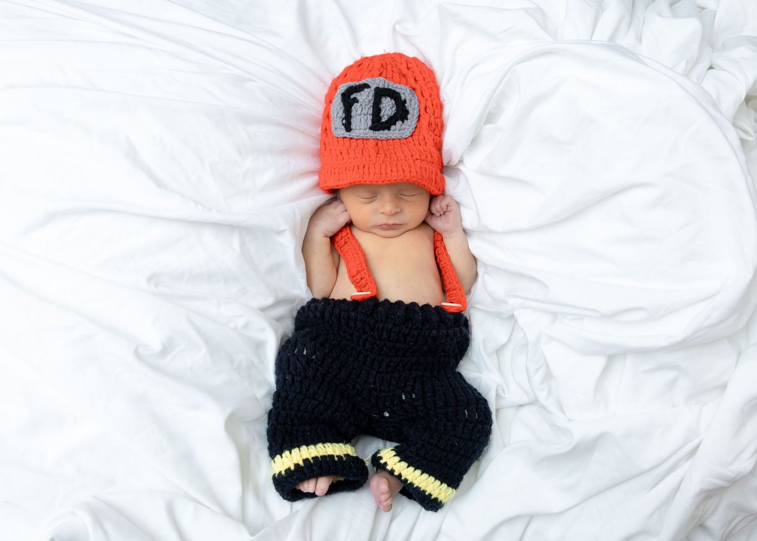 Adorable newborn baby dressed in a crocheted firefighter costume lying on white blanket.
by Andrew Fein Photography - Family and Newborn Pho