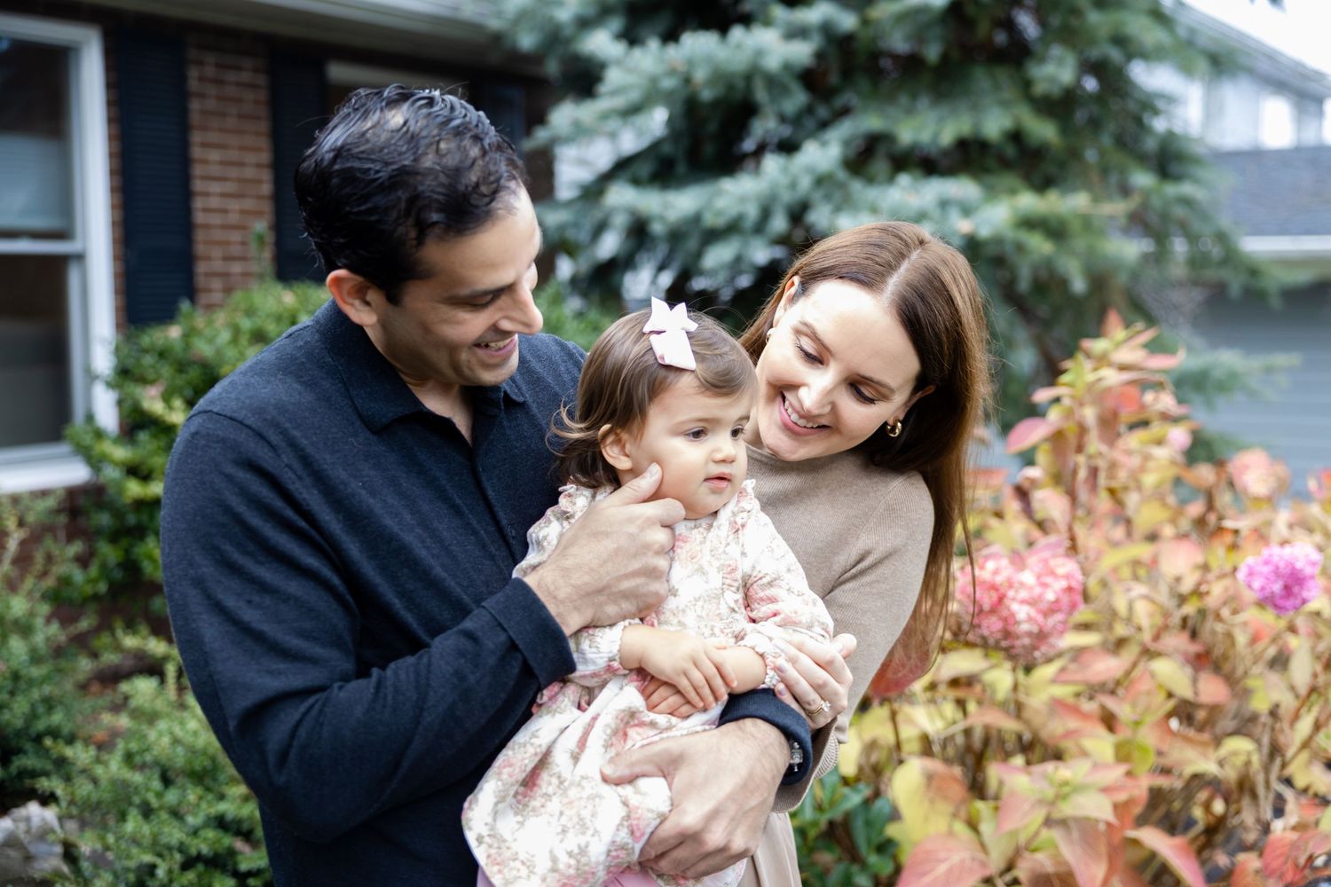 Happy young family with father holding baby girl smiling outdoors near garden flowers and brick house.