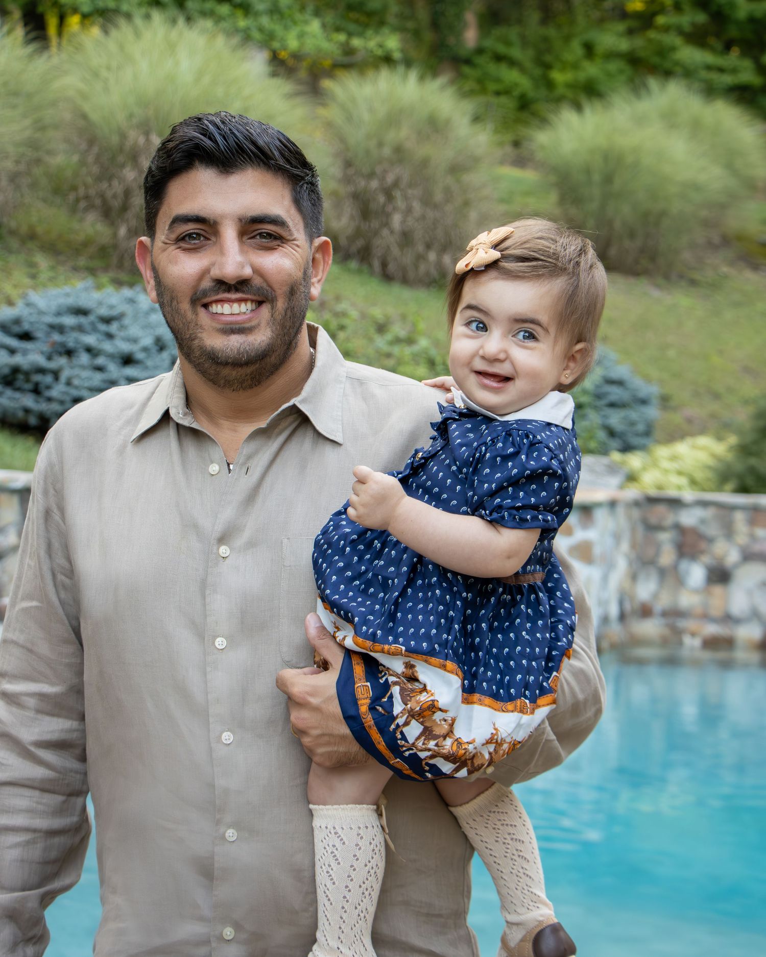 A smiling man holds a happy baby girl in a blue dress near a pool with lush greenery in the background.