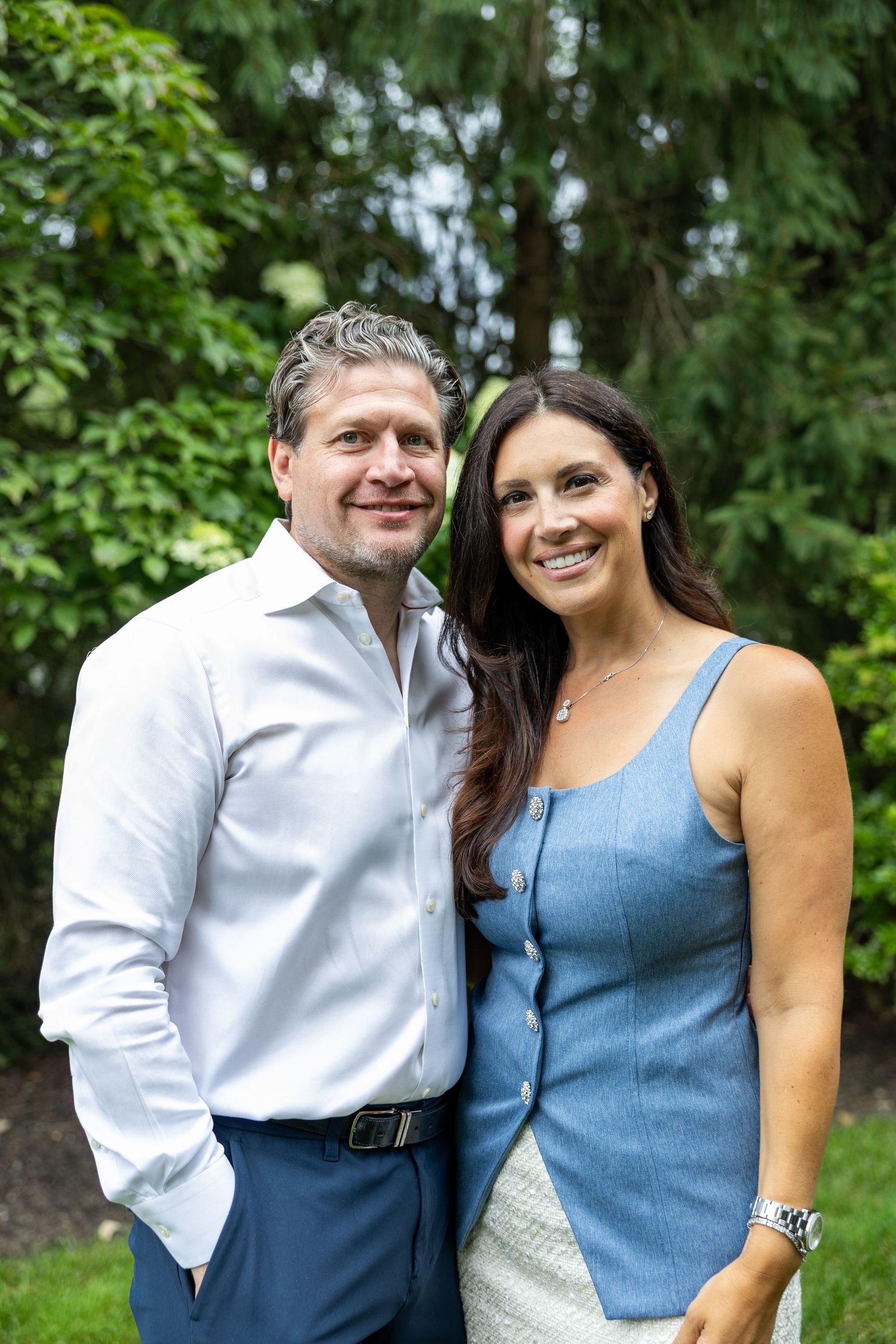 Smiling couple posing outdoors in a lush green garden, man in white shirt and woman in blue dress.