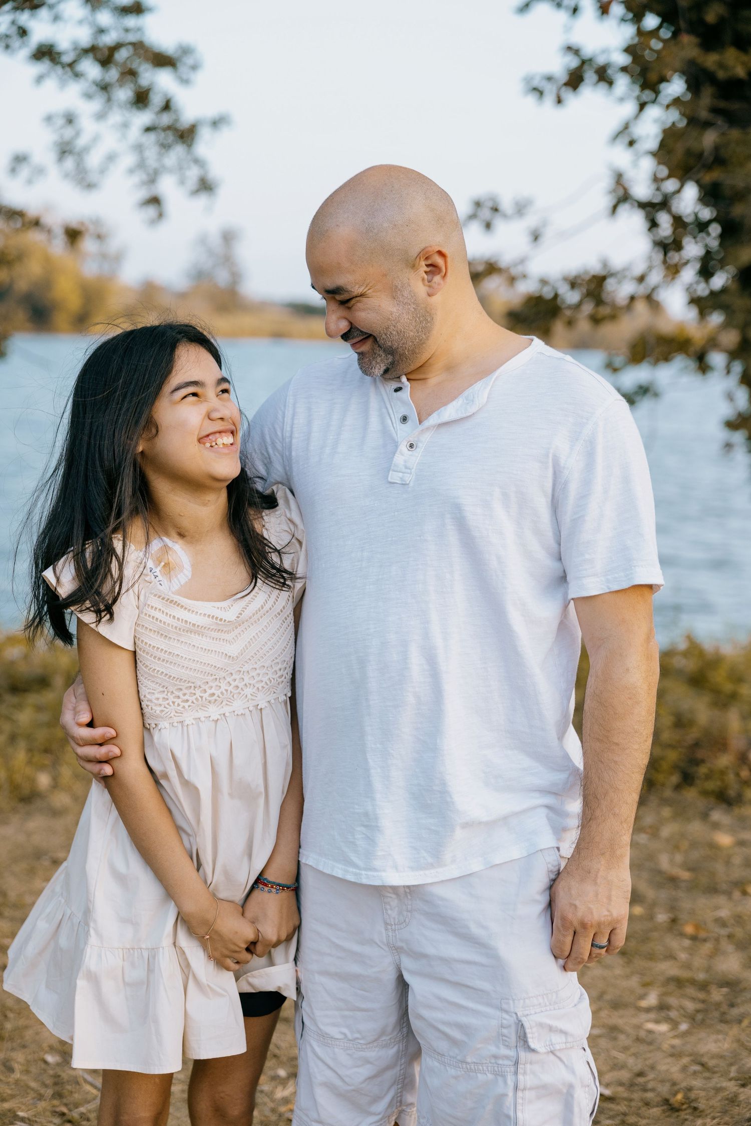 Bald man and young girl smiling at each other outdoors near a river, both wearing white.
by Andrew Fein Photography - Family and Newborn Pho