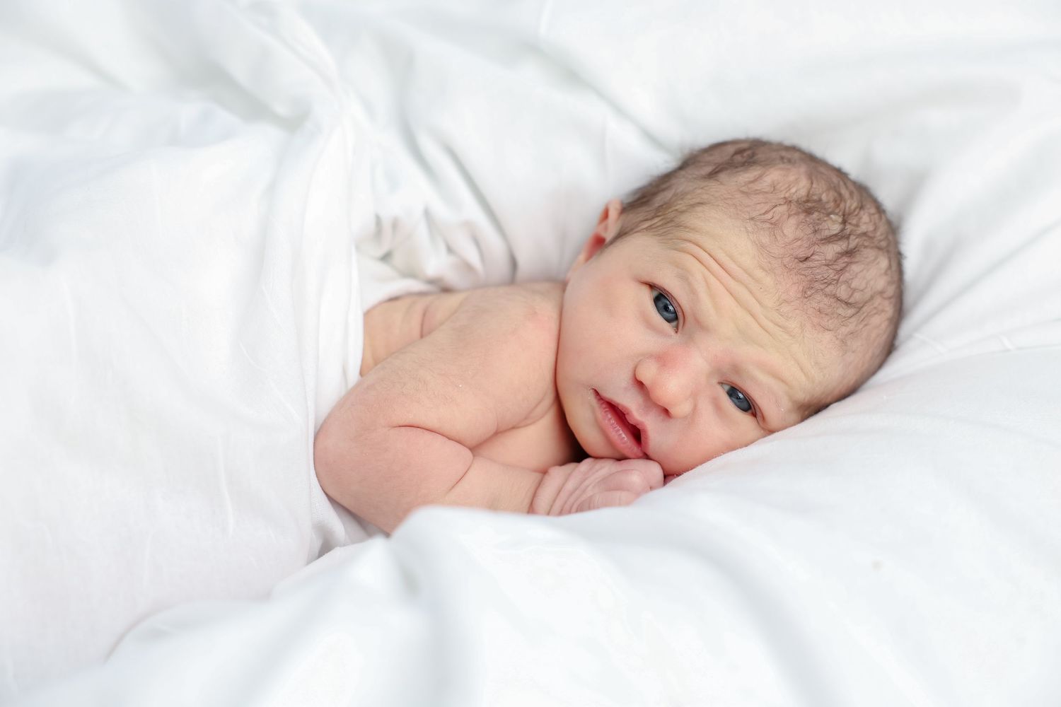 Newborn baby lying on white blankets, looking peacefully at the camera with blue eyes.