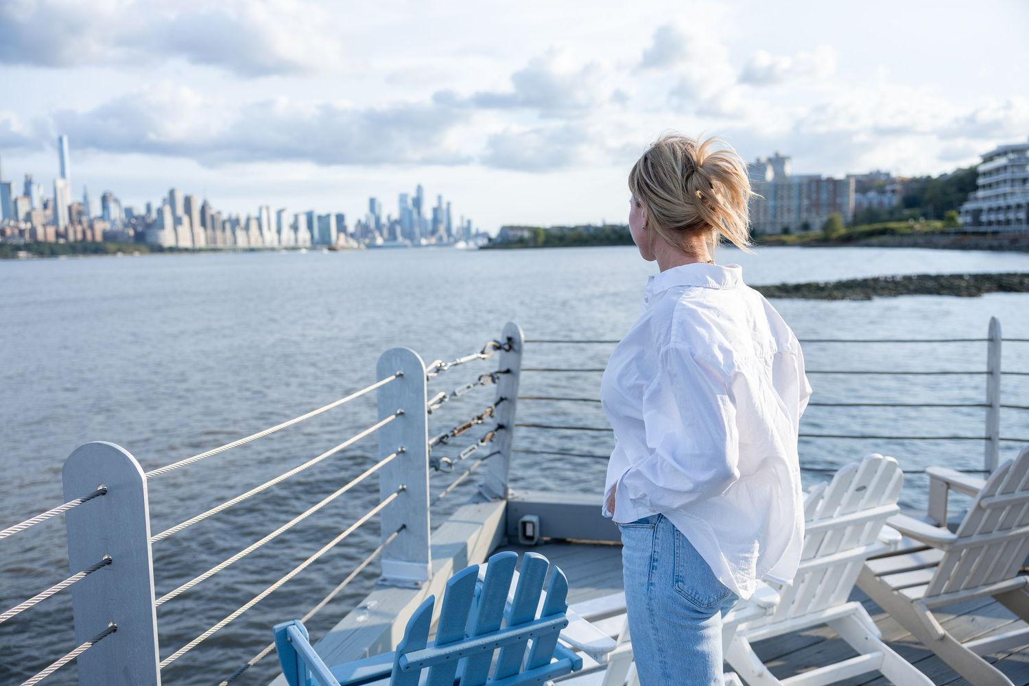 Woman in white shirt gazes at NYC skyline from a waterfront deck on a cloudy day.