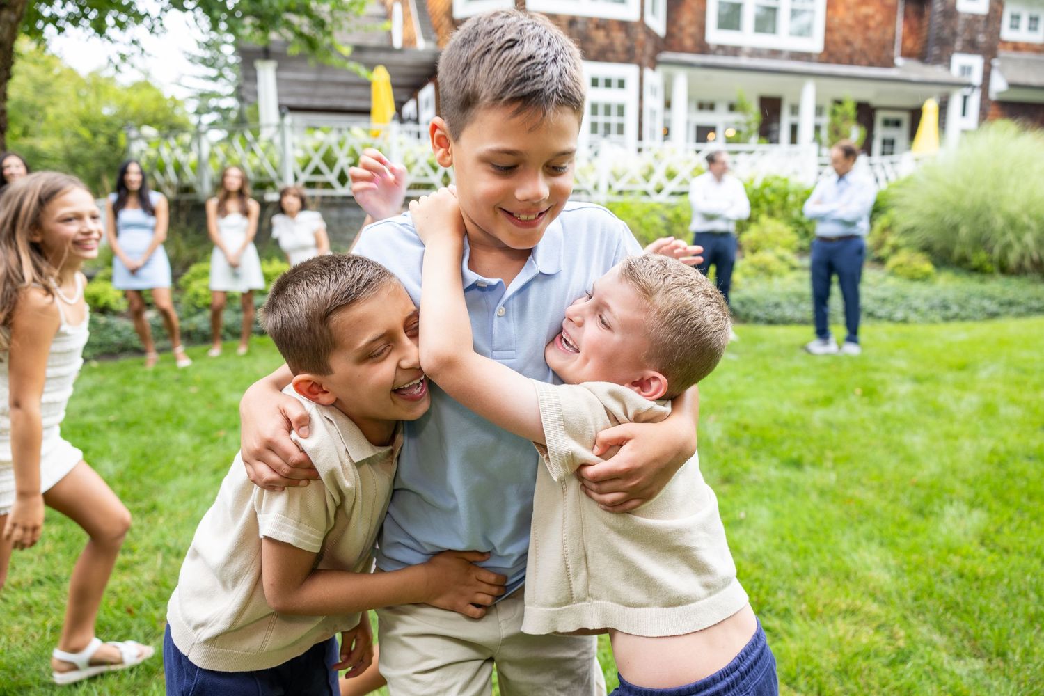 Three boys hugging and laughing together outdoors at a garden party with other children in the background.
