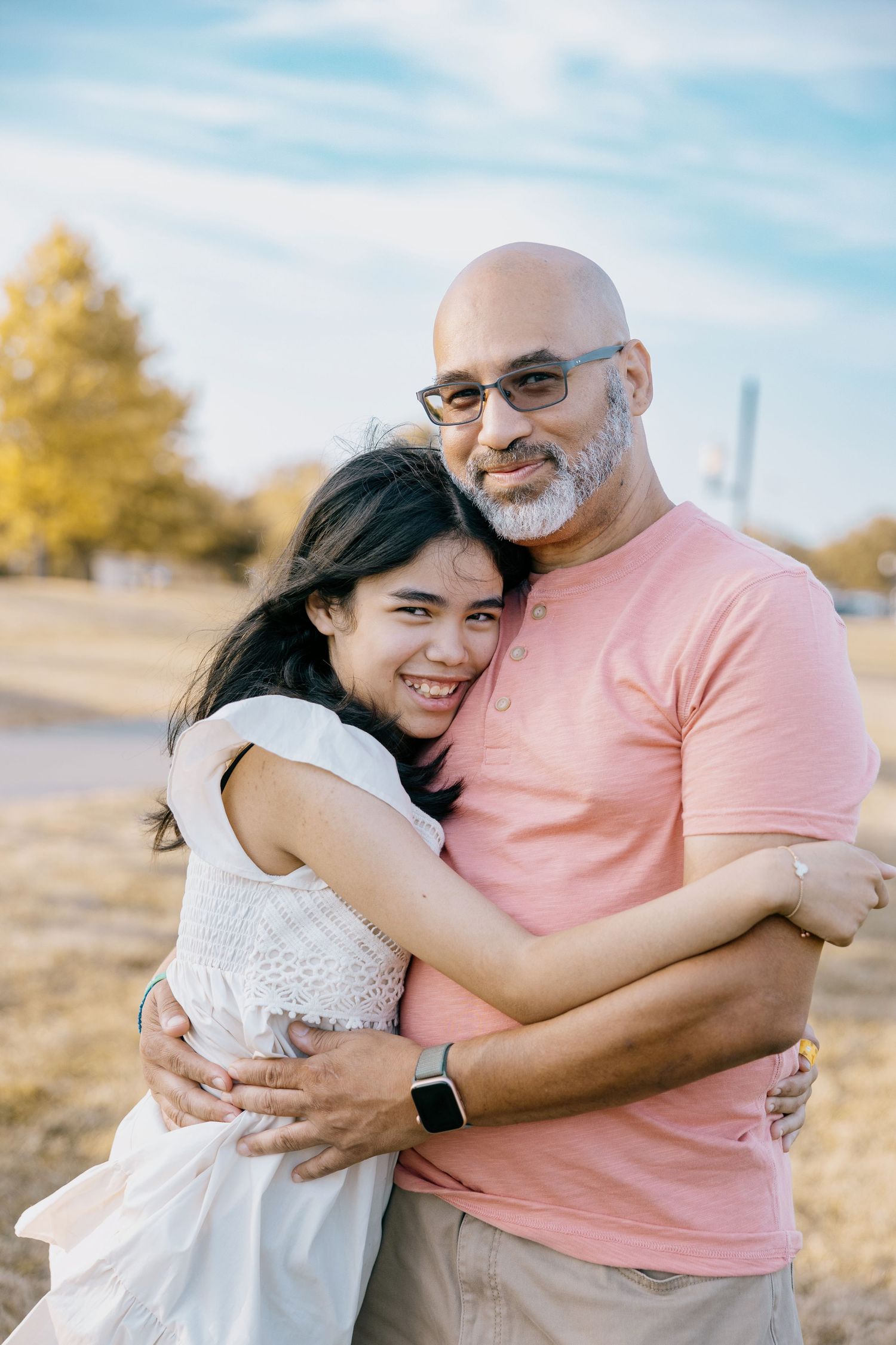 A smiling couple embraces outdoors, with a bald man in a pink shirt and a woman in a white dress, set against a blue sky.