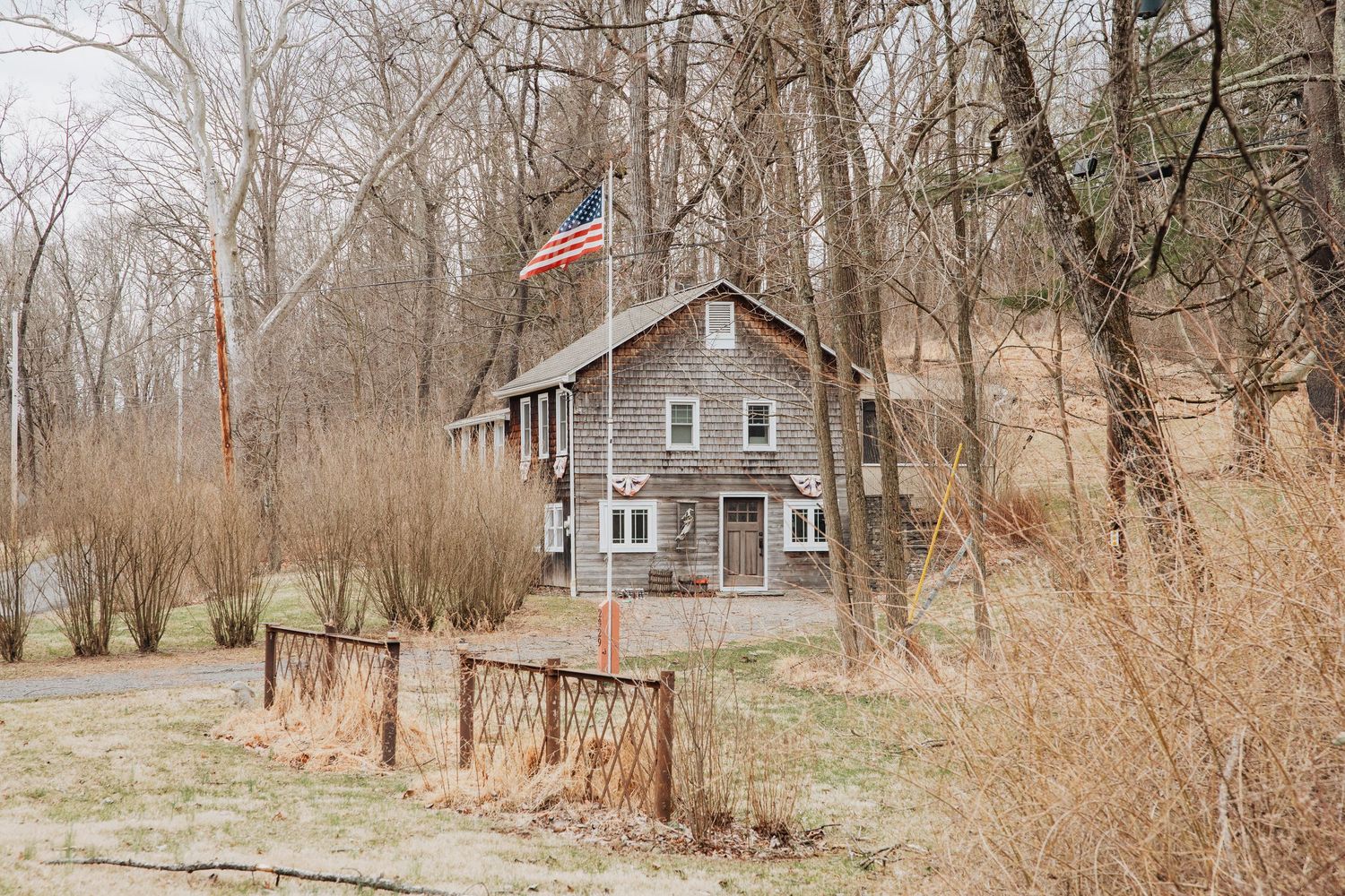 Rustic wooden cabin surrounded by bare trees with an American flag flying proudly in a wooded rural setting.