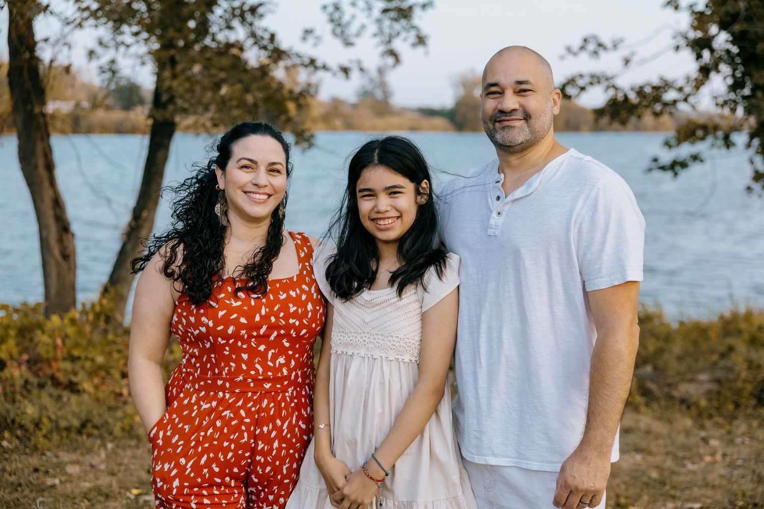 Family of three smiling outdoors near a lake, woman in red dress, girl in white dress, man in white shirt.