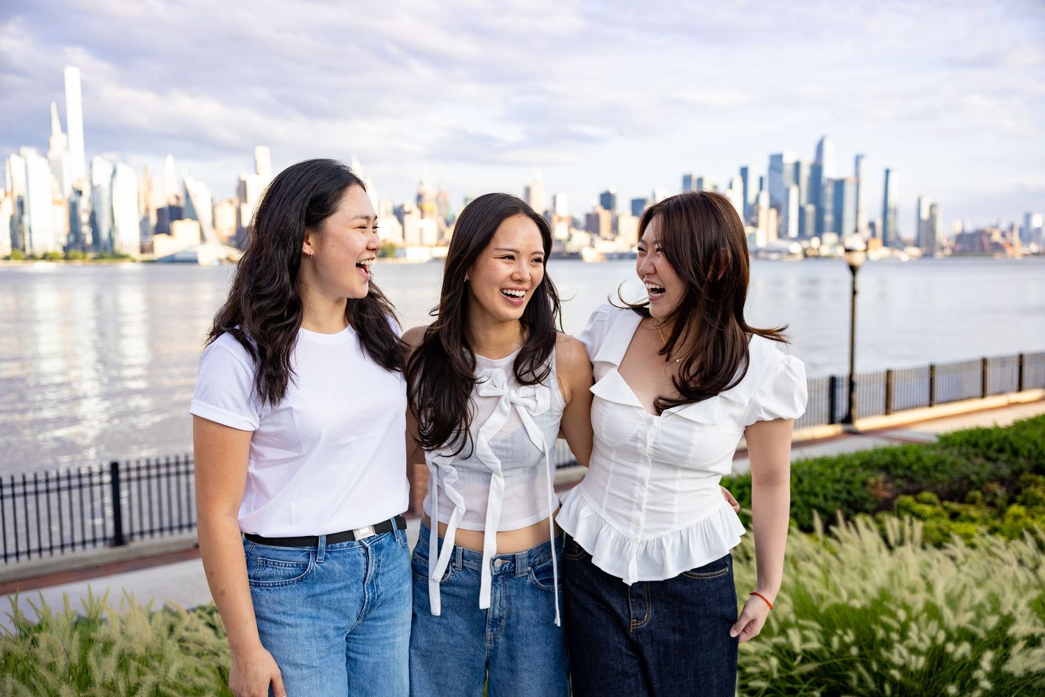 Three young women laughing together outdoors with NYC skyline and Hudson River in the background.