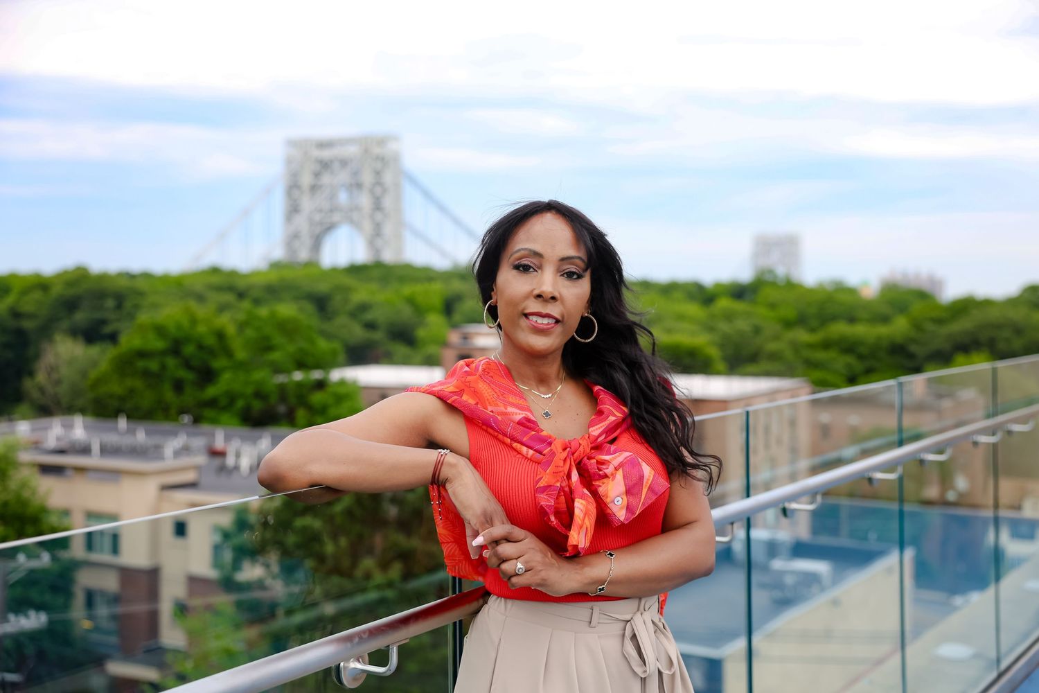Woman in red top poses on rooftop terrace with George Washington Bridge and lush trees in background.