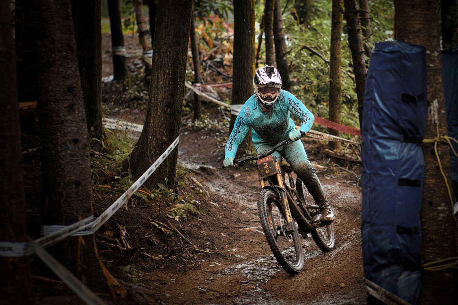 Racing downhill through muddy forest trail wearing teal jersey and white helmet by Dave Clément, Québec.